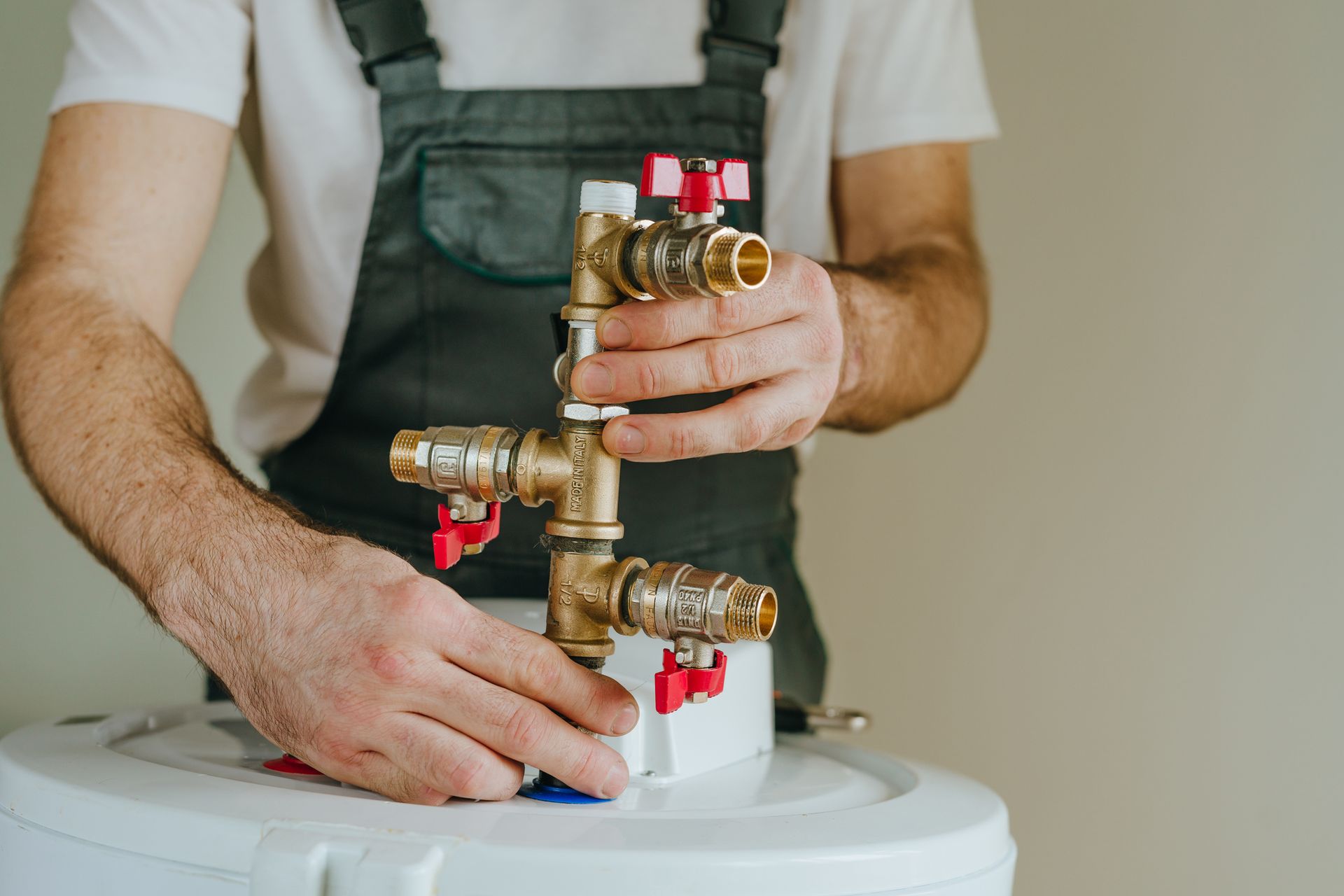 Plumber in overalls connecting brass pipes and valves to a white water heater.