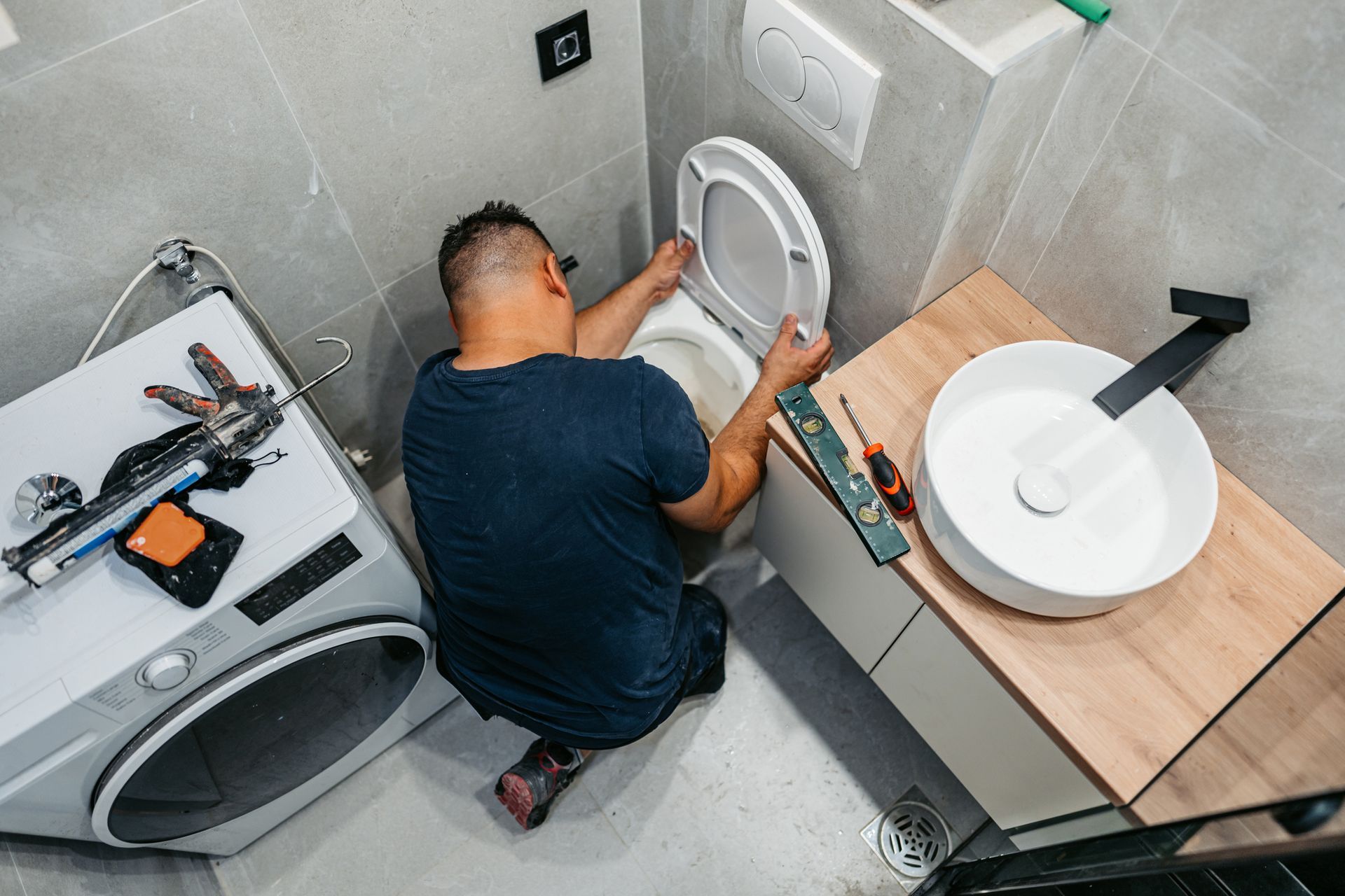 Man installing a toilet seat in a modern bathroom with a sink, washing machine, and tools.