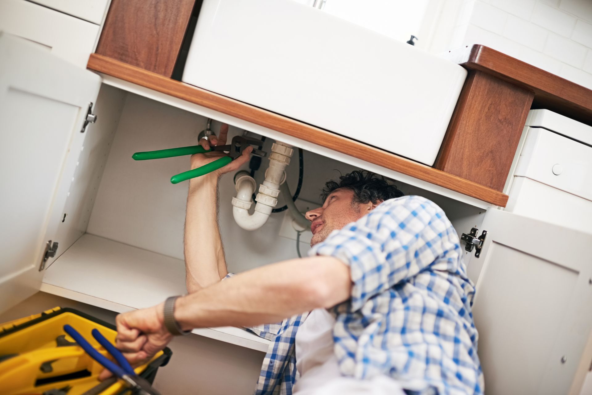 Person with curly hair, in a plaid shirt, works on plumbing under a kitchen sink using pliers.
