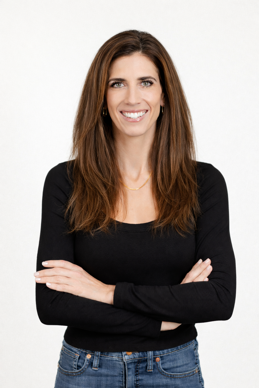 A woman in a black shirt and blue jeans is standing with her arms crossed and smiling.