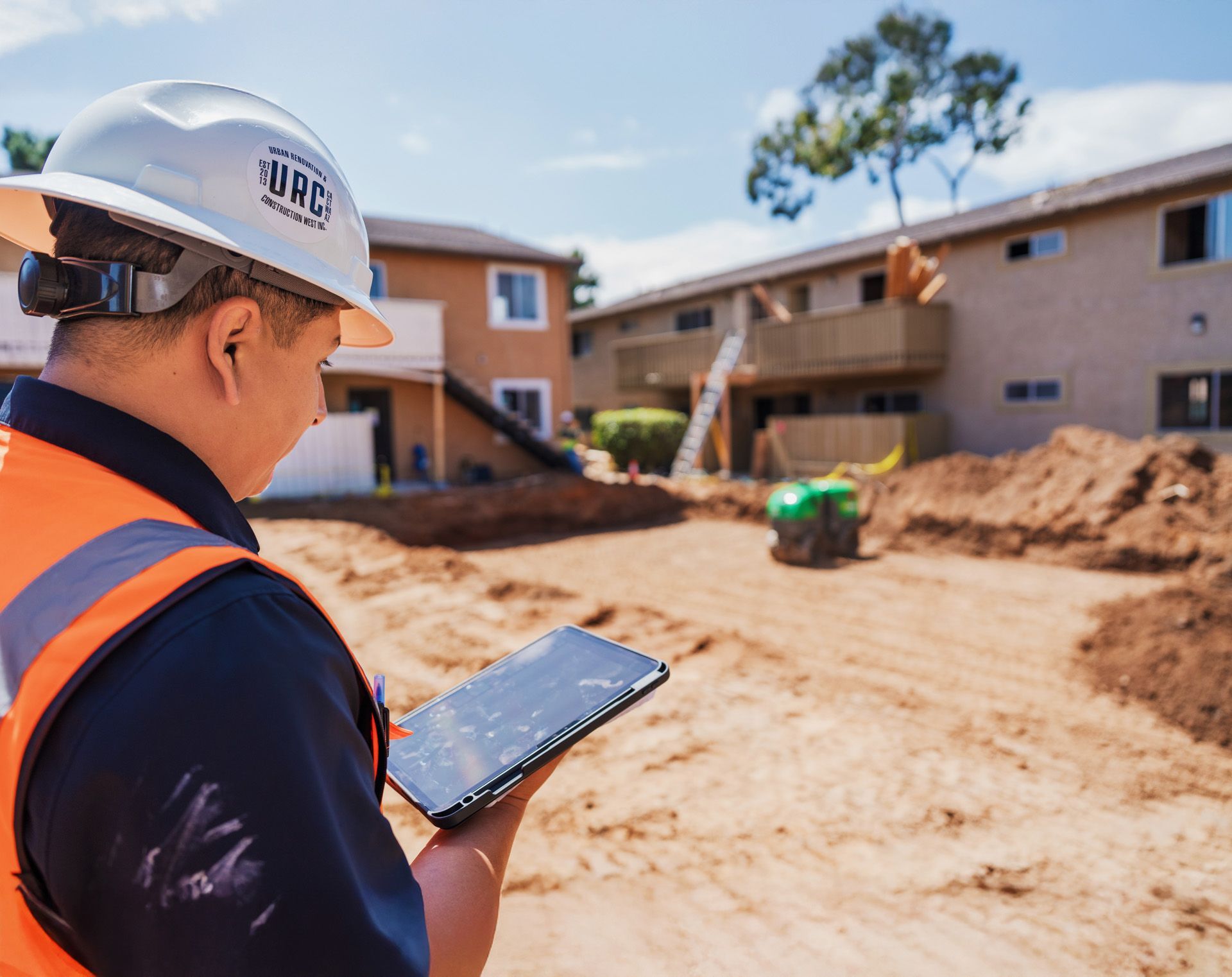 Construction worker in hard hat using a tablet at a building site with dirt piles and houses in the background