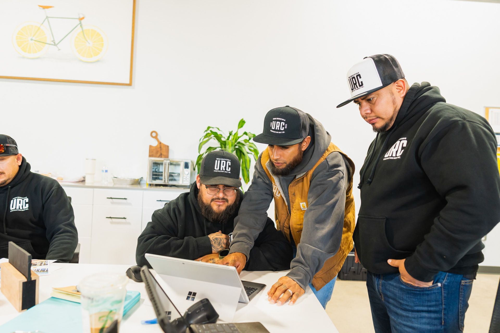 Three men in caps review a laptop in a bright office, leaning over a desk.
