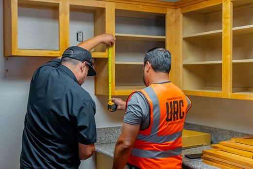 Two workers installing kitchen cabinets, one in an orange safety vest measuring a countertop area.