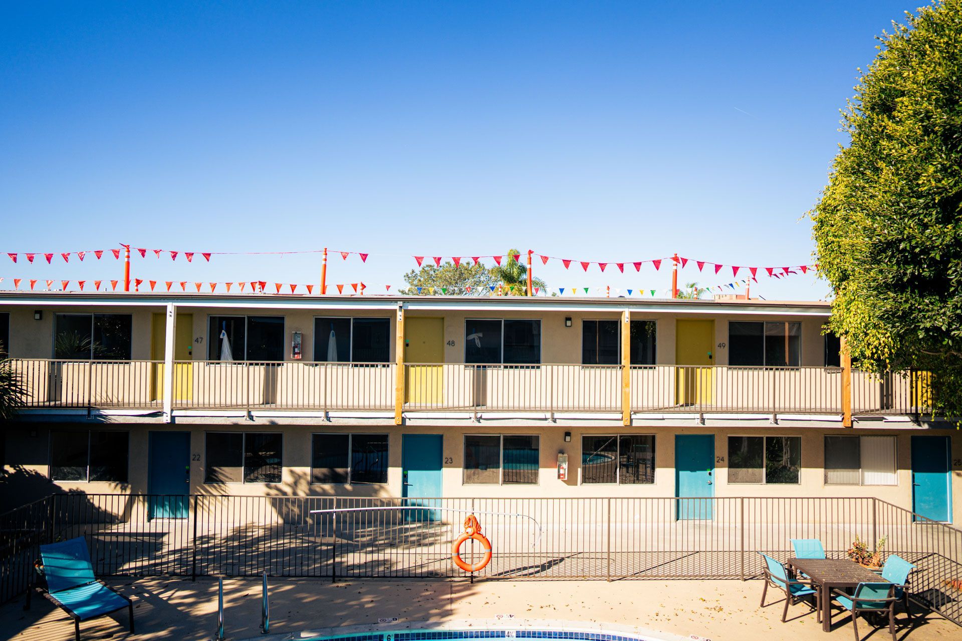 Motel-style building with balconies, red bunting, and a poolside patio under a clear blue sky