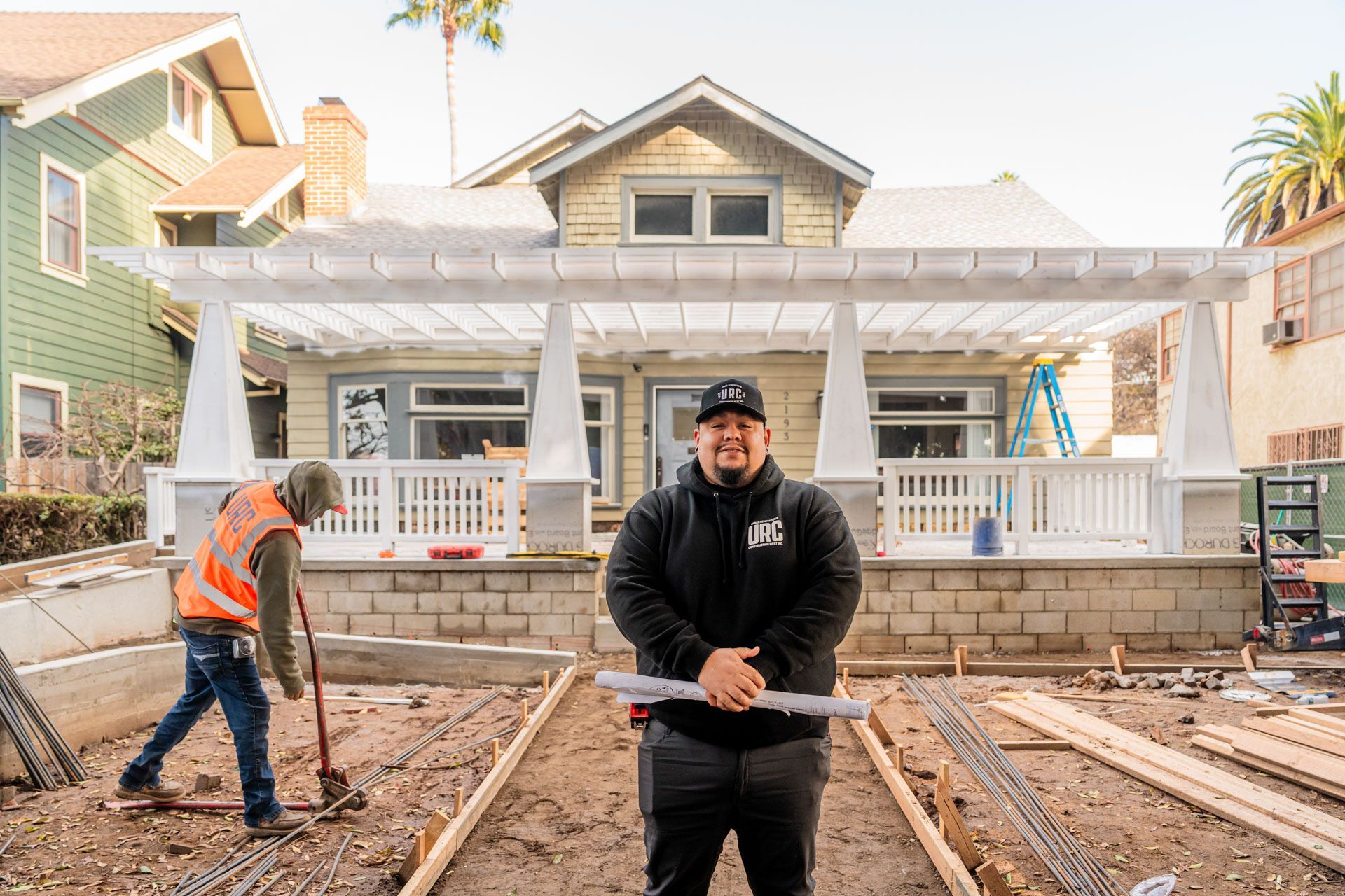 Construction workers at a house renovation site, one standing in front of the porch and one working with tools.