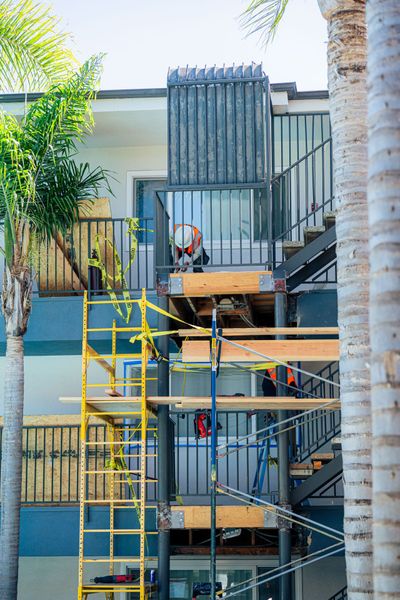 Scaffold and stairs outside a building with workers and yellow safety rails, framed by palm trees