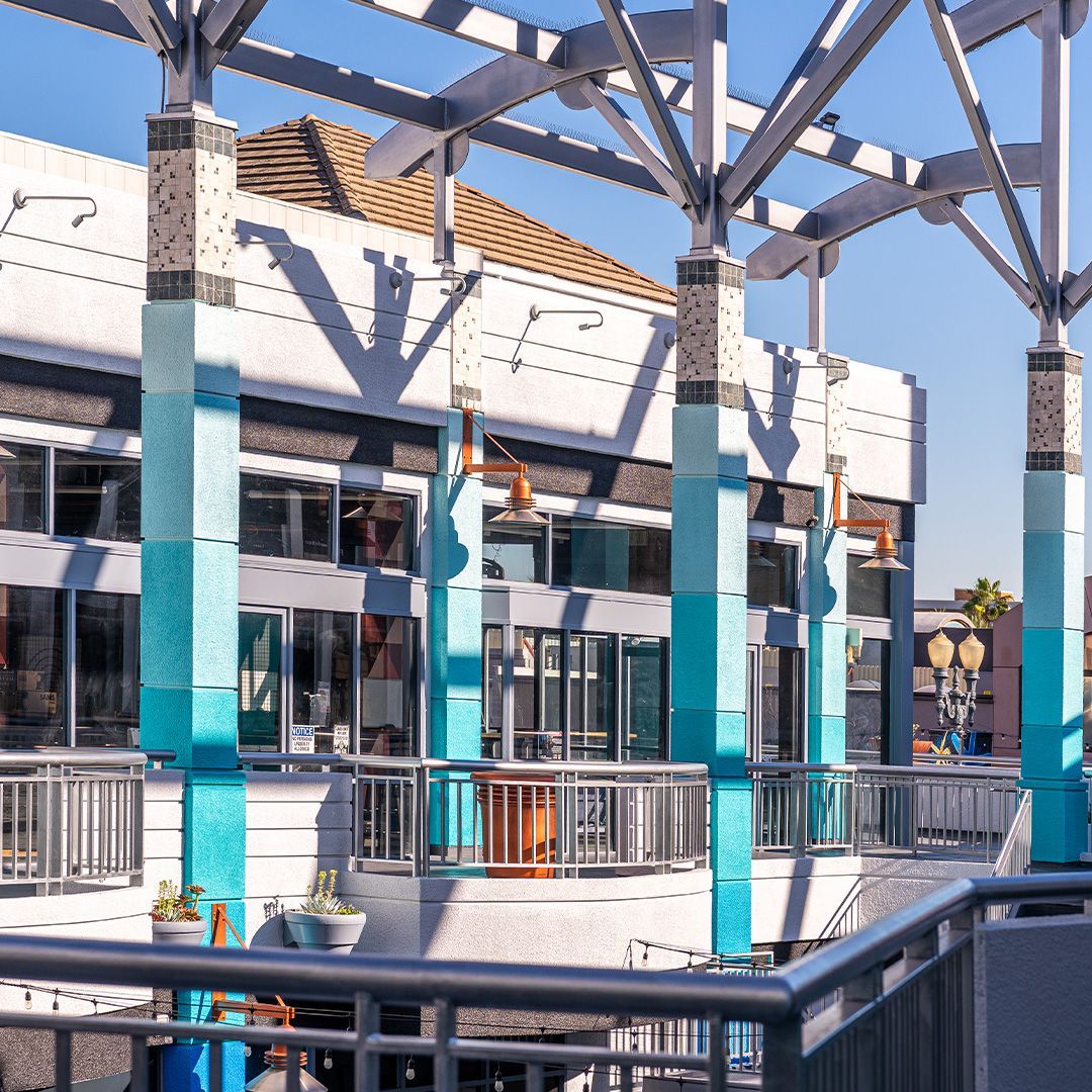 Outdoor shopping center with blue and white columns, metal roof and railings, sunny day.