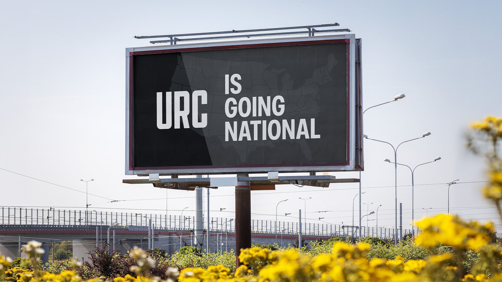 Billboard reading “URC is Going National” above yellow flowers by a roadside