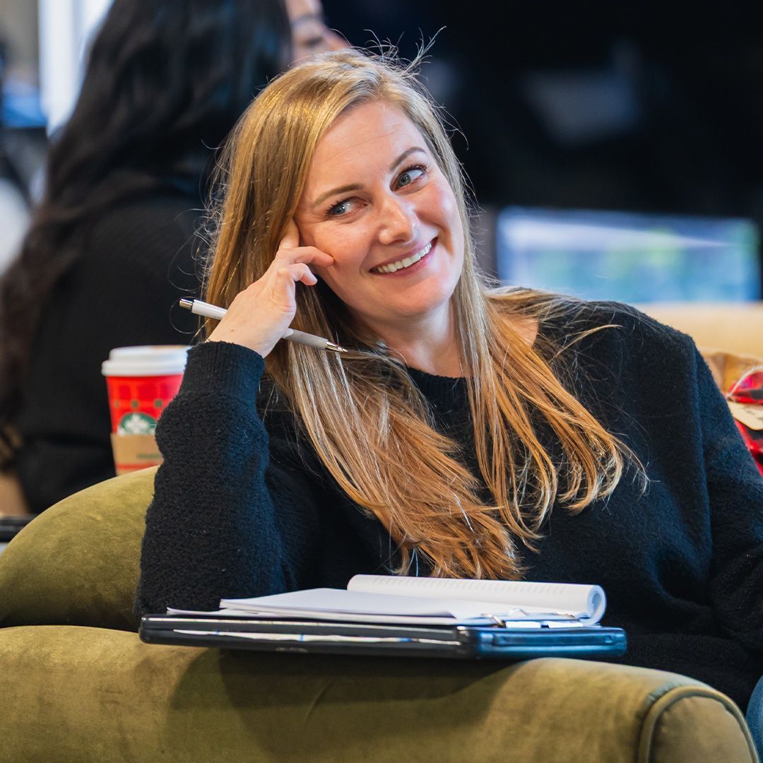 Woman with blonde hair smiles, resting elbow on sofa. Holds pen, notebook, and coffee cup nearby.