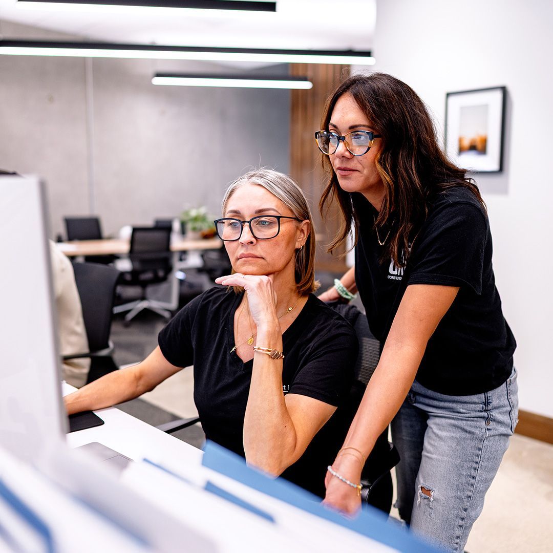 Two women in office looking at computer screen, one pointing, with papers and office background.