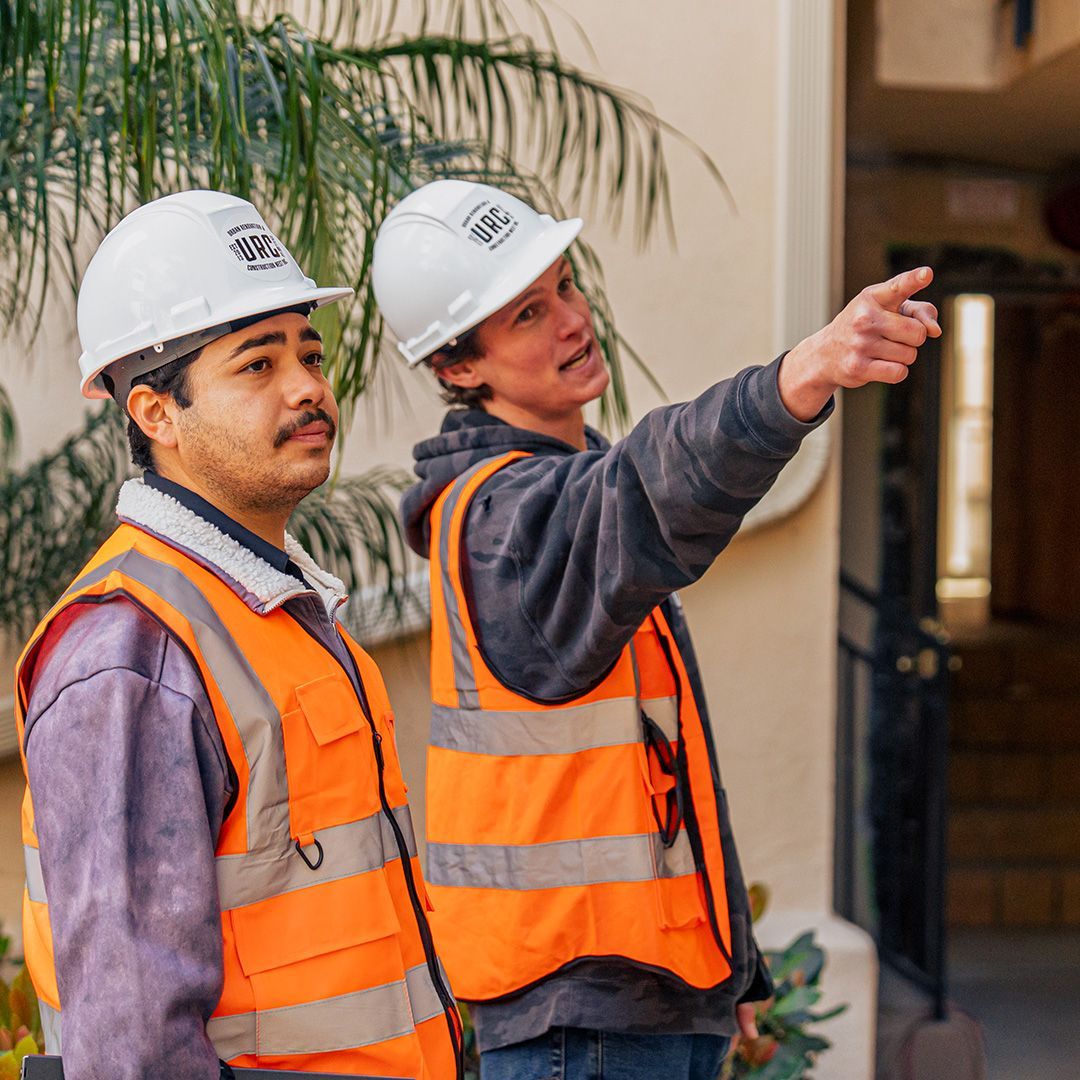 Two construction workers in hard hats and vests, one pointing upwards.