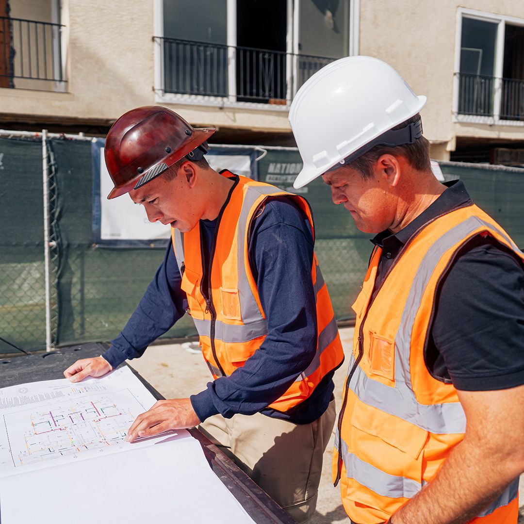 Two construction workers in safety vests and hard hats reviewing blueprints outdoors.