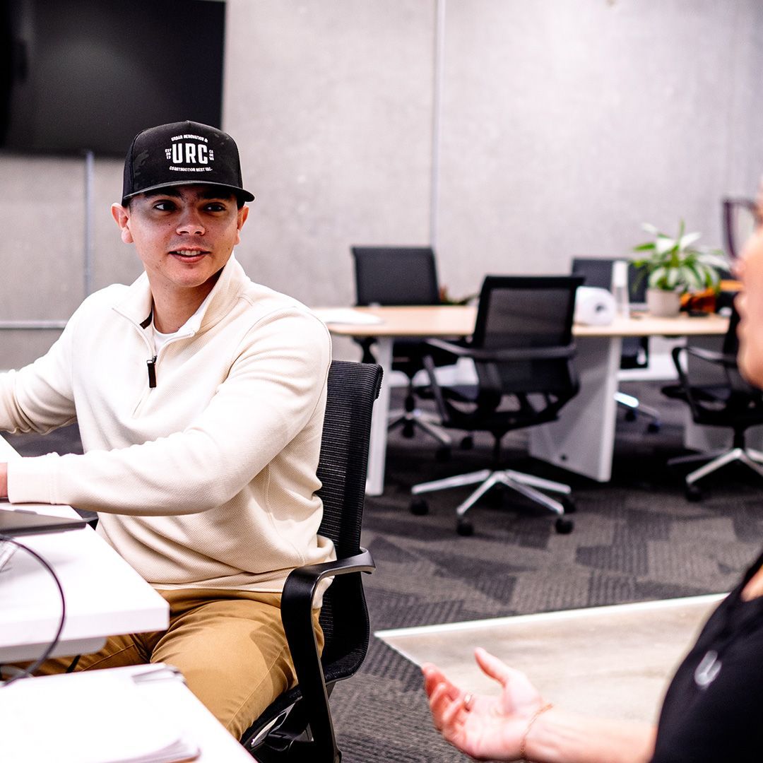 Man in a beige sweater and cap talking to another person in an office setting.