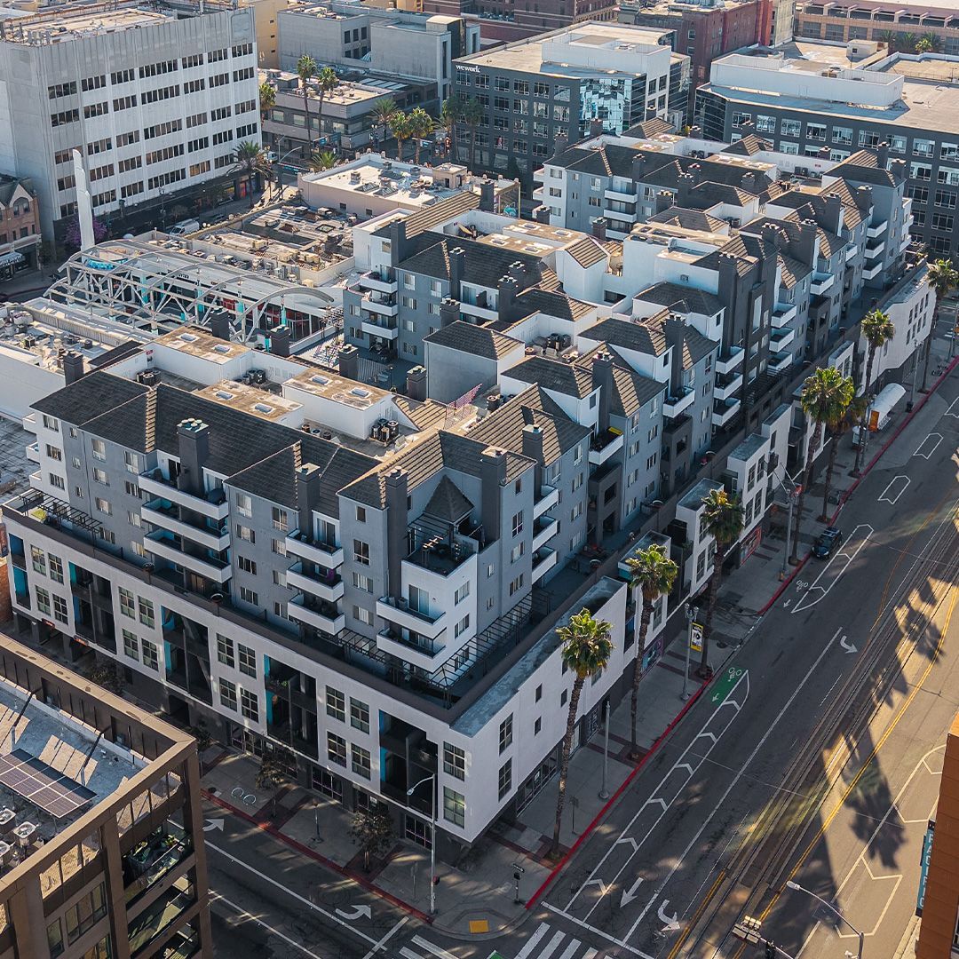 Aerial view of a multi-story apartment building with a white and gray facade, palm trees, and a city street.