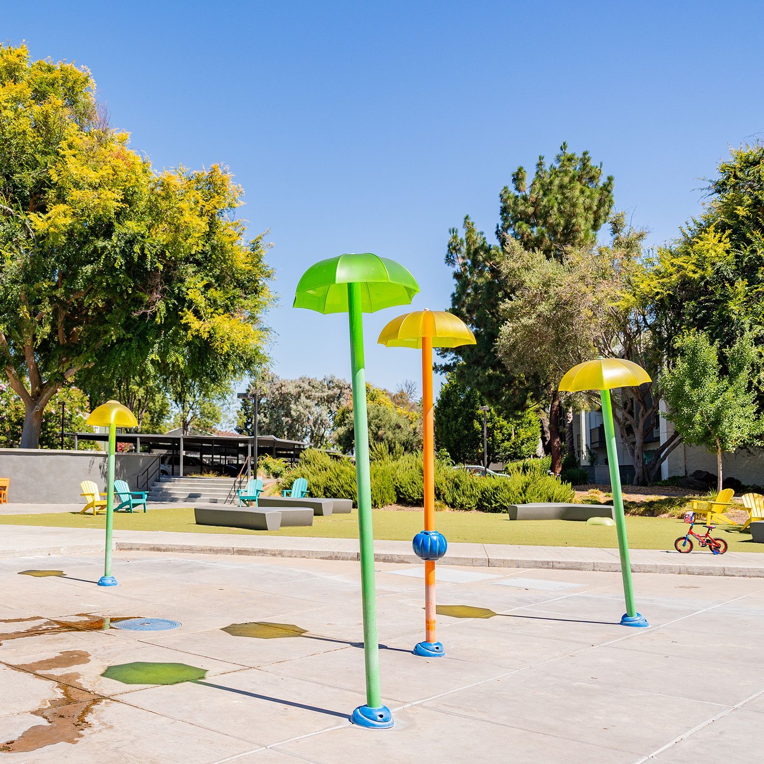 Playground splash pad with colorful umbrella-shaped water features. Green trees and clear blue sky in background.