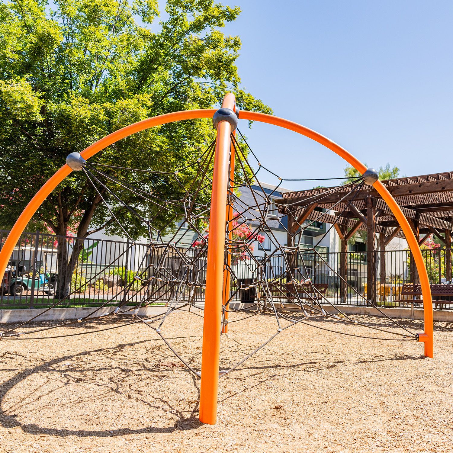 Orange climbing structure with rope netting in a playground, against a backdrop of a tree and wooden pergola.