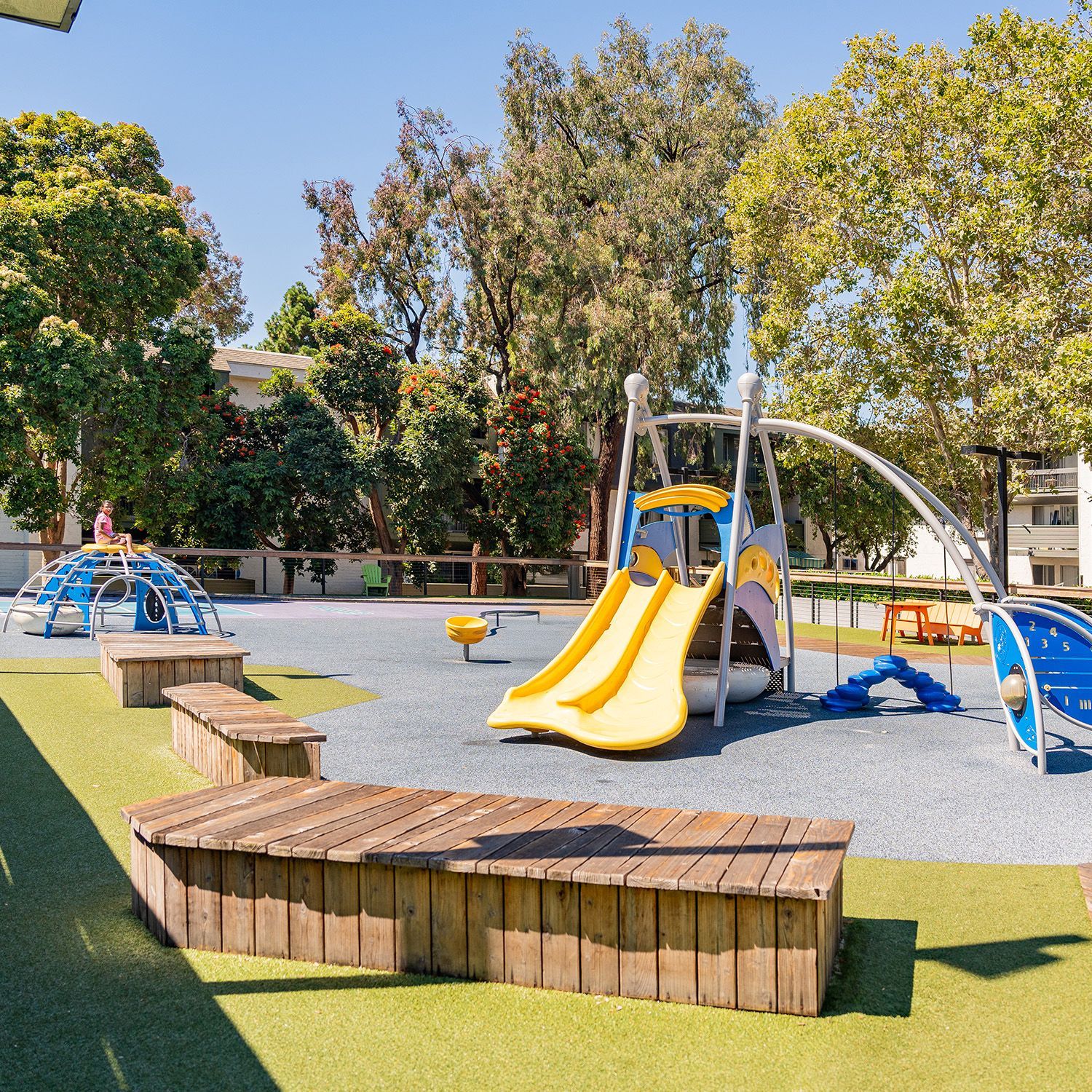 Playground with a yellow slide, benches, and play structures. Green grass and trees in the background.