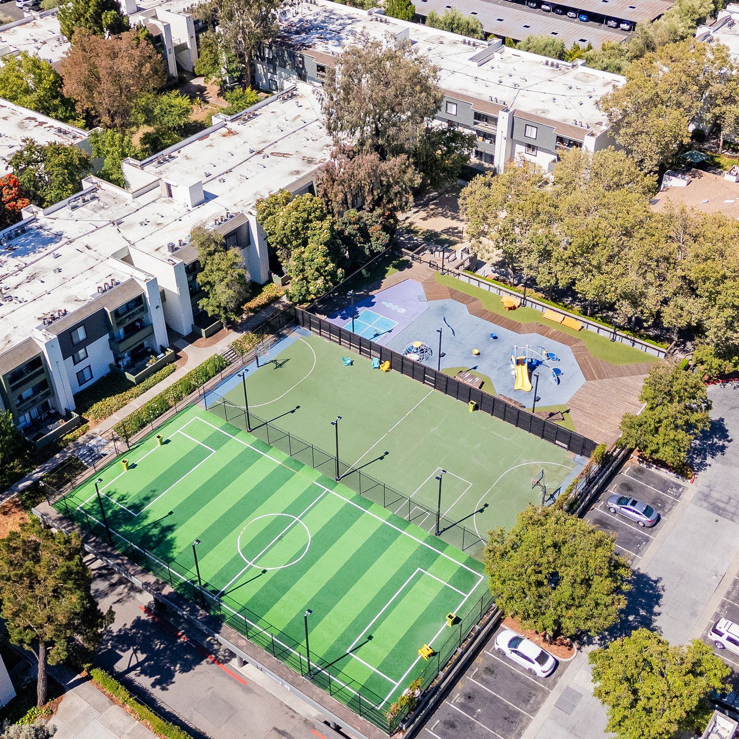 Aerial view of a sports complex with a soccer field, playground, parking, and apartments.