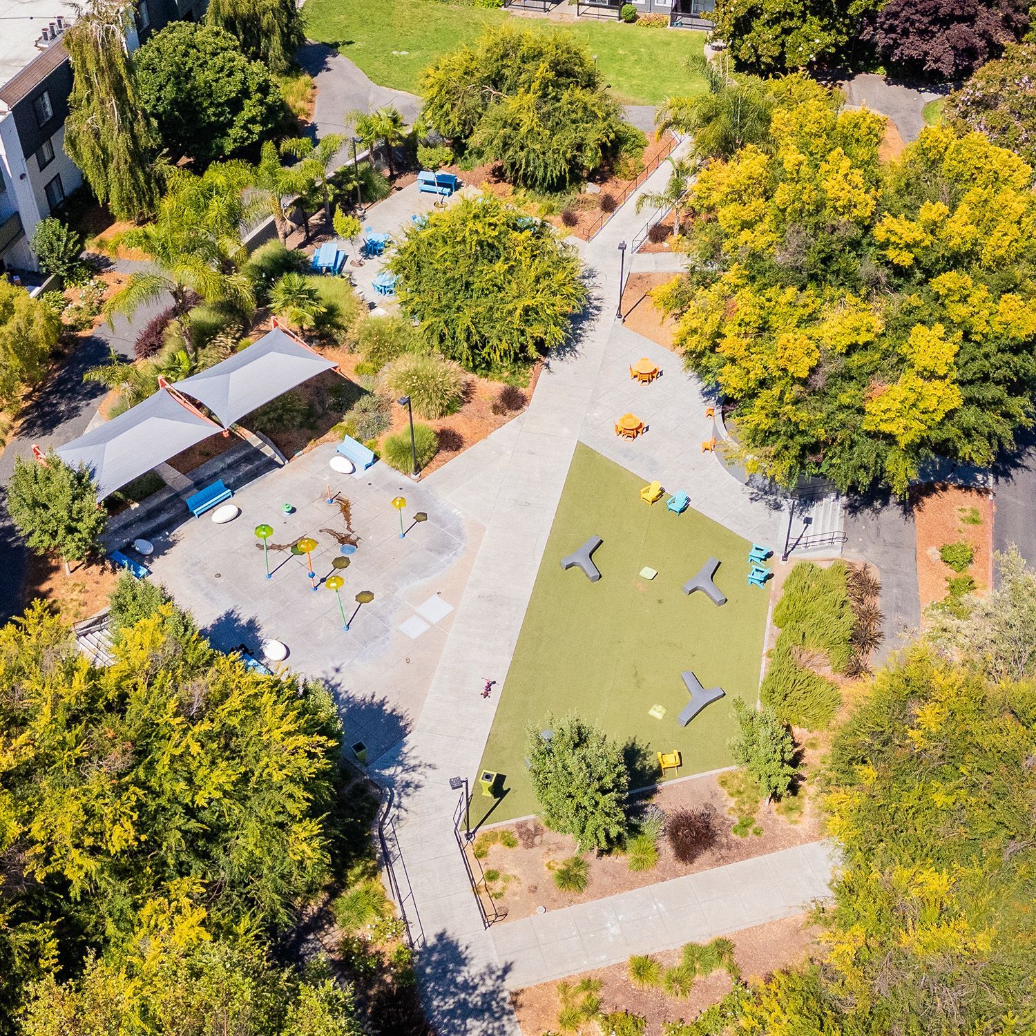 Aerial view of a playground with green and tan play areas, shaded seating, and surrounding trees.