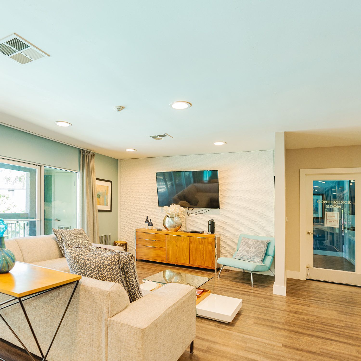 Living room with beige couch, TV on a wooden cabinet, teal accent chair, and glass coffee table.