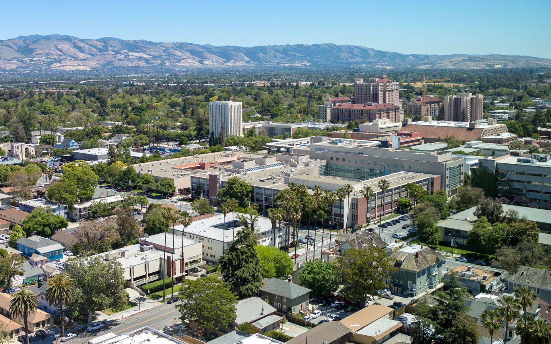 Aerial view of San Jose, California, buildings and trees under blue sky, mountains in background.