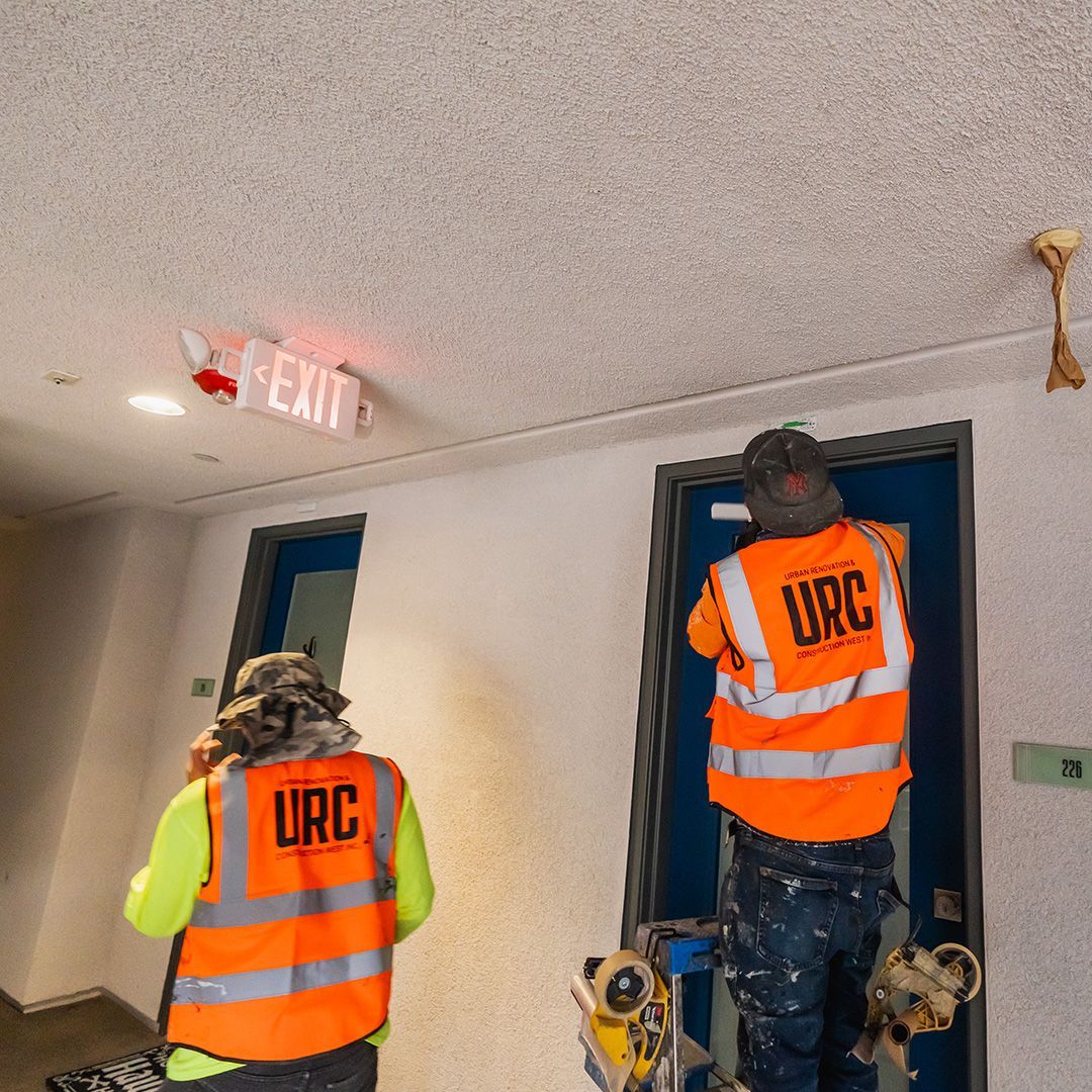 Two workers in orange vests are renovating an interior hallway. One works on a door, the other on the wall.