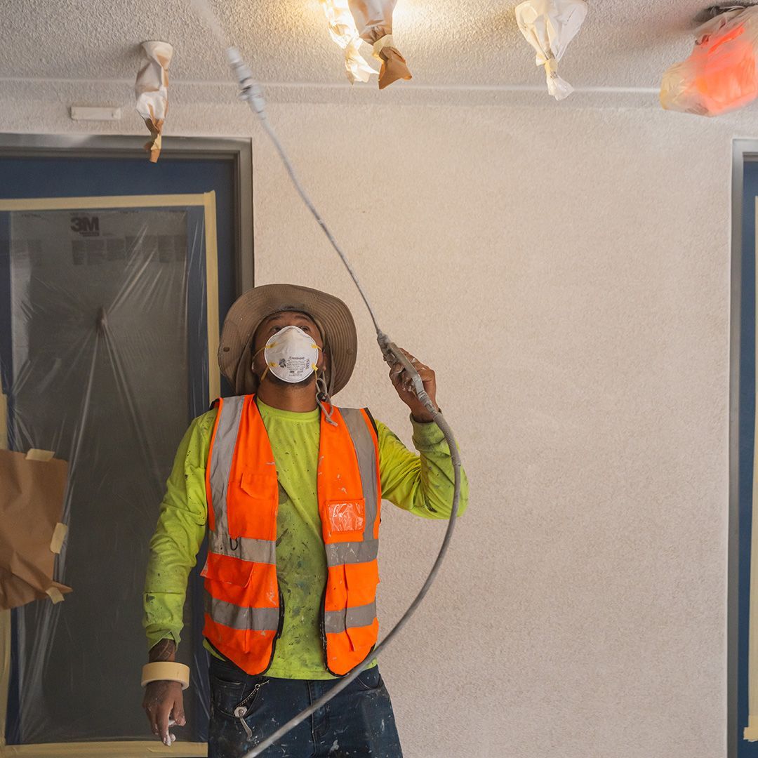 Construction worker spraying ceiling with a long spray gun, wearing a safety vest, mask, and hat.