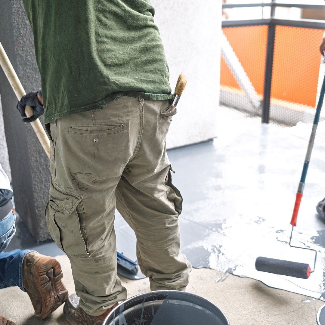 Person painting a floor with a roller. They wear a green shirt and tan cargo pants. A paintbrush sticks out from a pocket.
