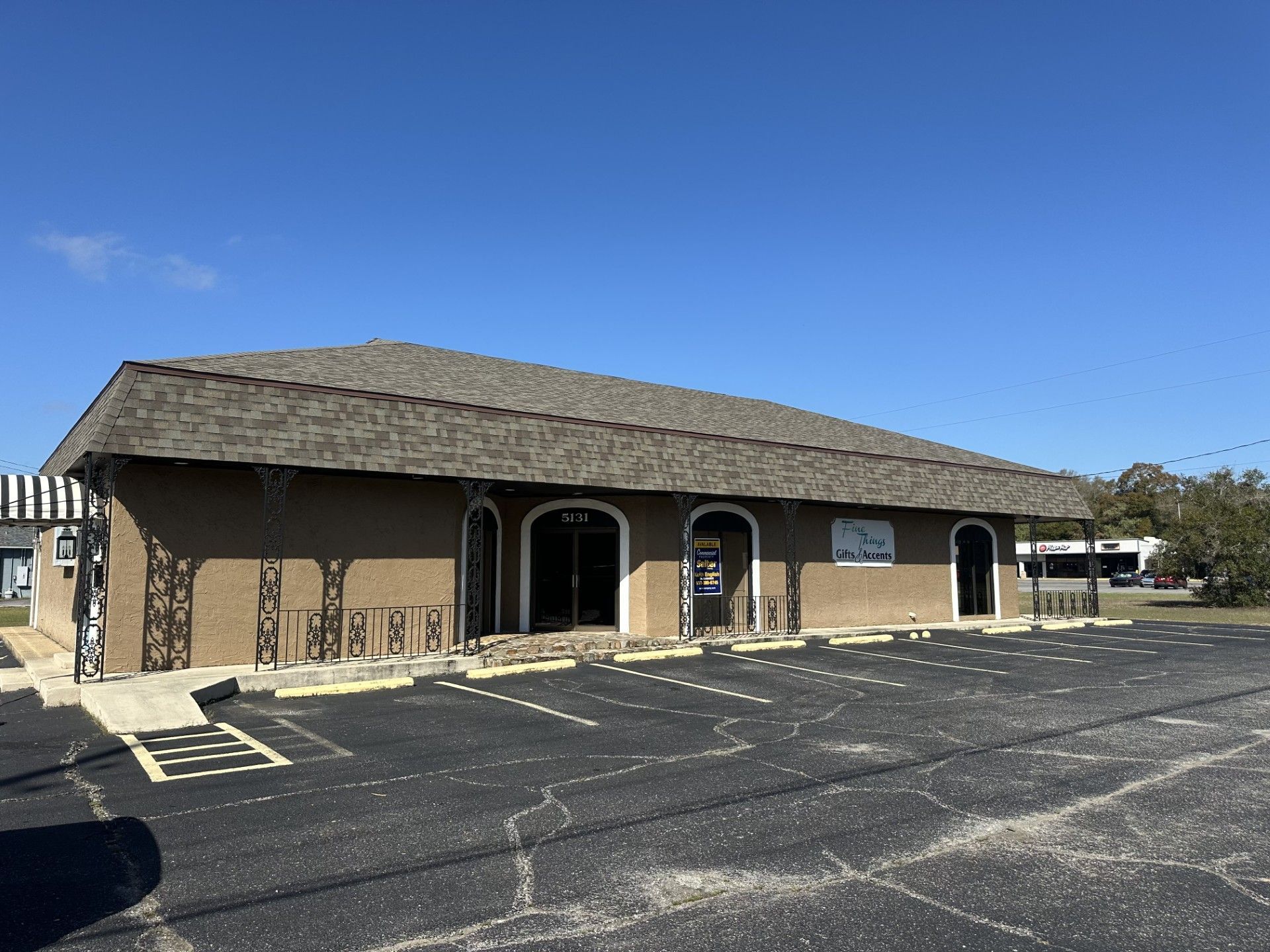 A single-story, tan-colored commercial building with a grey shingled roof, arched doorways, and a large empty parking lot.