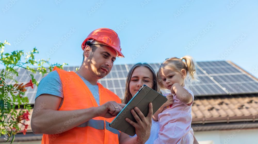 Man in orange vest and hardhat shows tablet to woman and child in front of solar panels.
