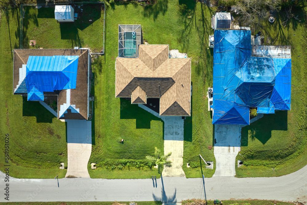 Aerial view of three houses with driveways, grass, and a street. Two houses have blue tarp roofs.