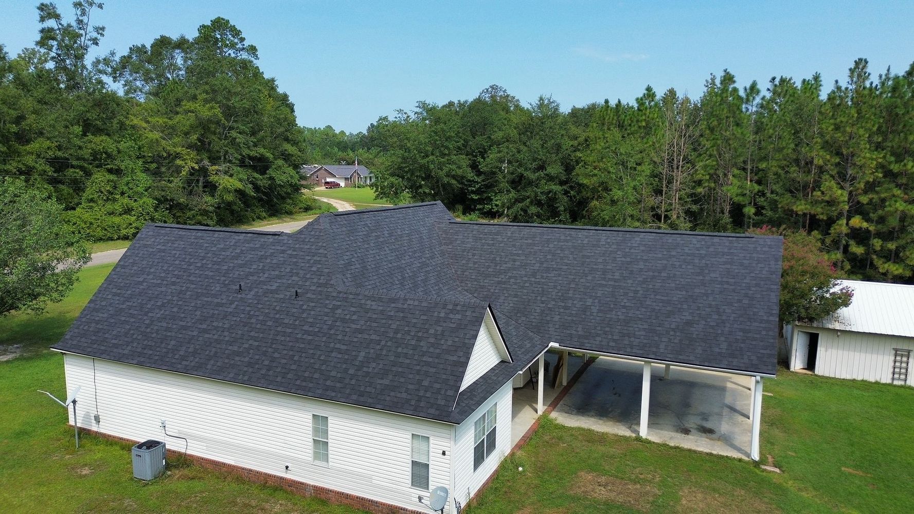 Aerial view of a white house with a dark roof and carport, set in a grassy area with trees under a blue sky.