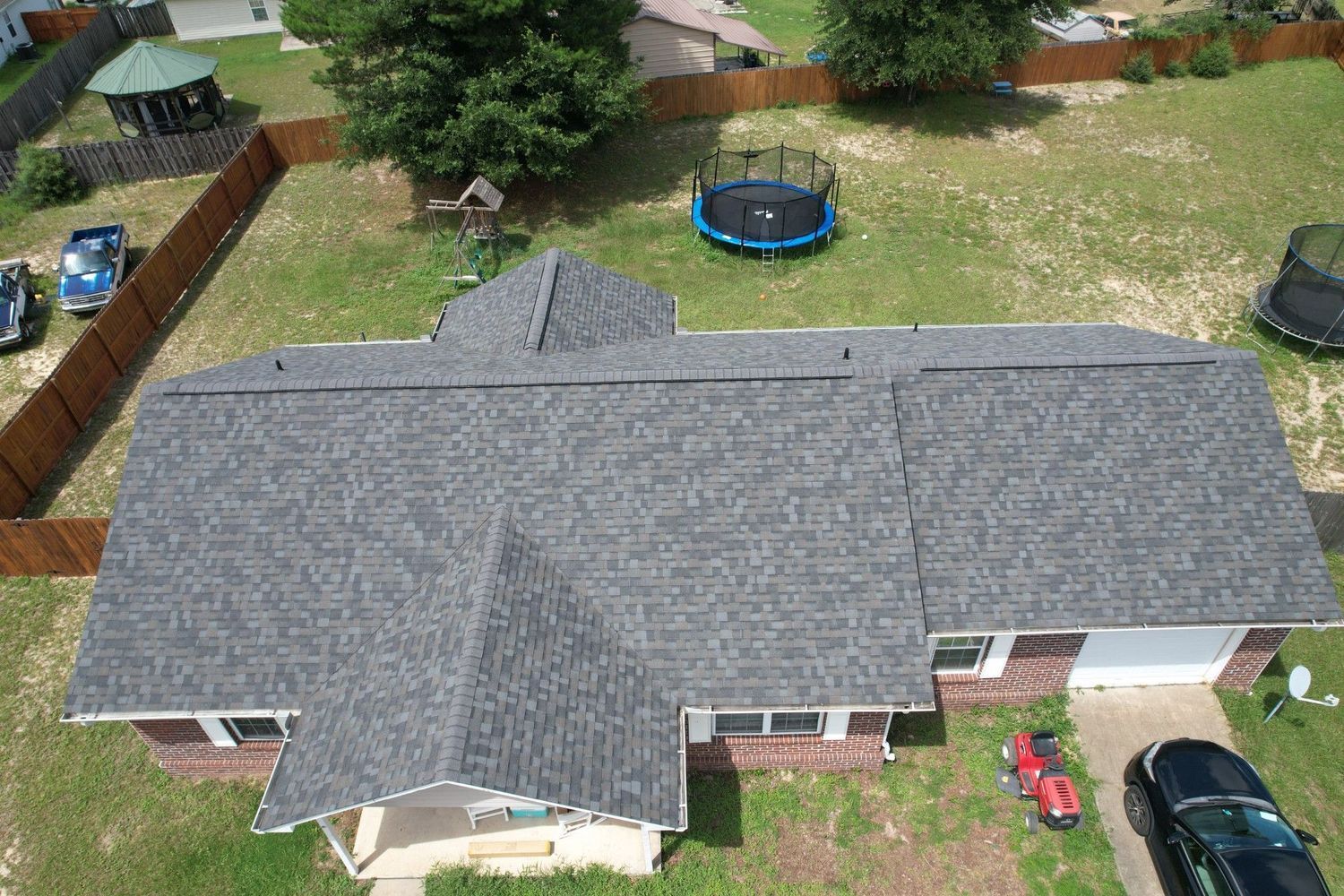 Aerial view of a house with gray shingle roof, surrounded by a yard with a trampoline, and a fence.