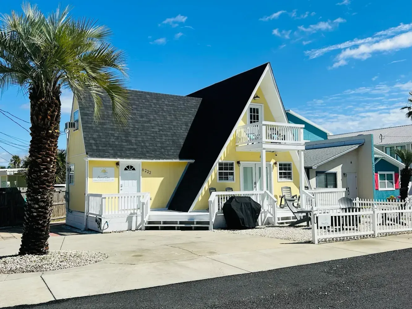 Yellow A-frame house with black roof, white deck, and palm tree. Sunny day.