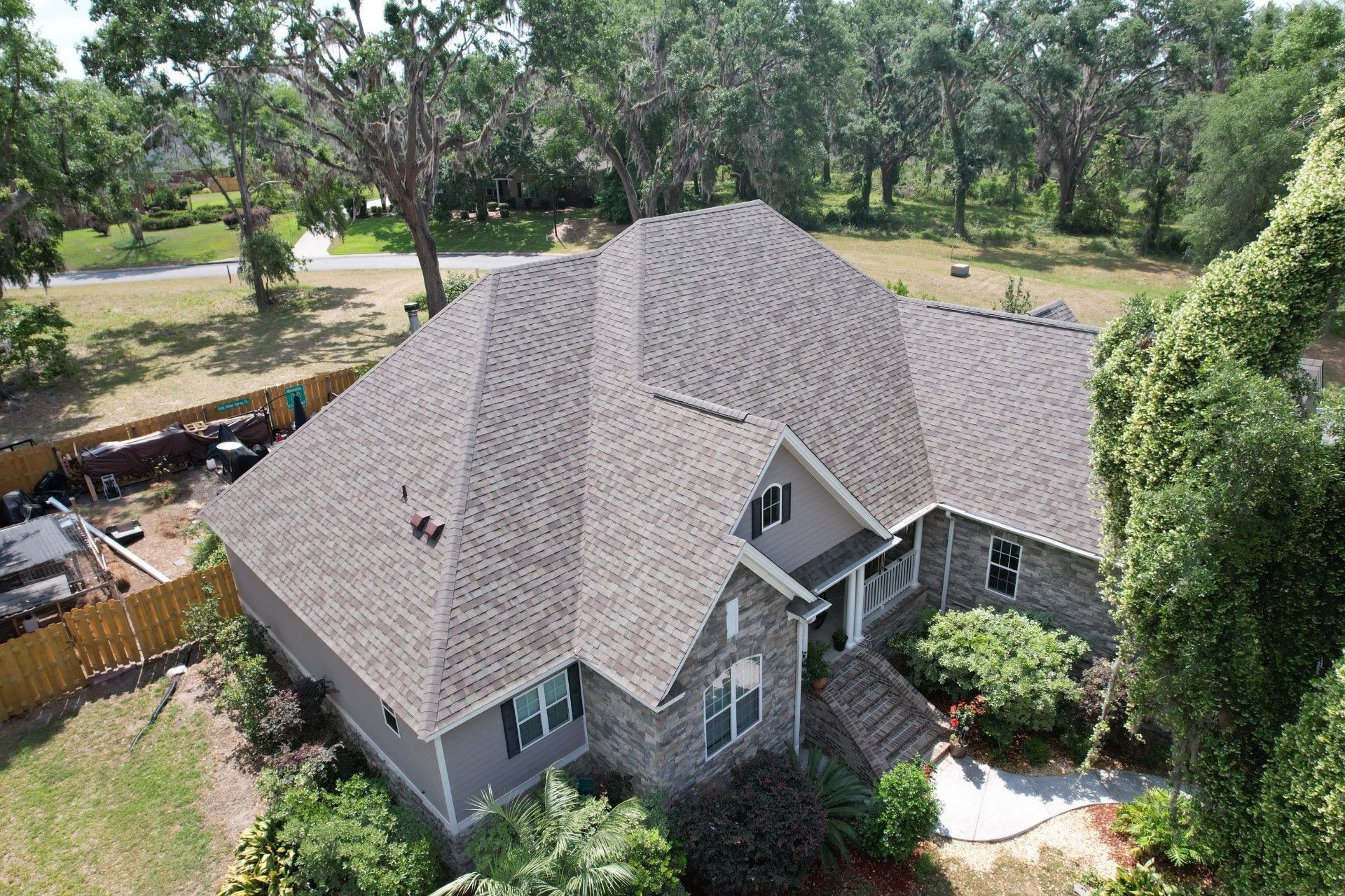 Aerial view of a house with a gray roof and siding, surrounded by trees and a wooden fence.