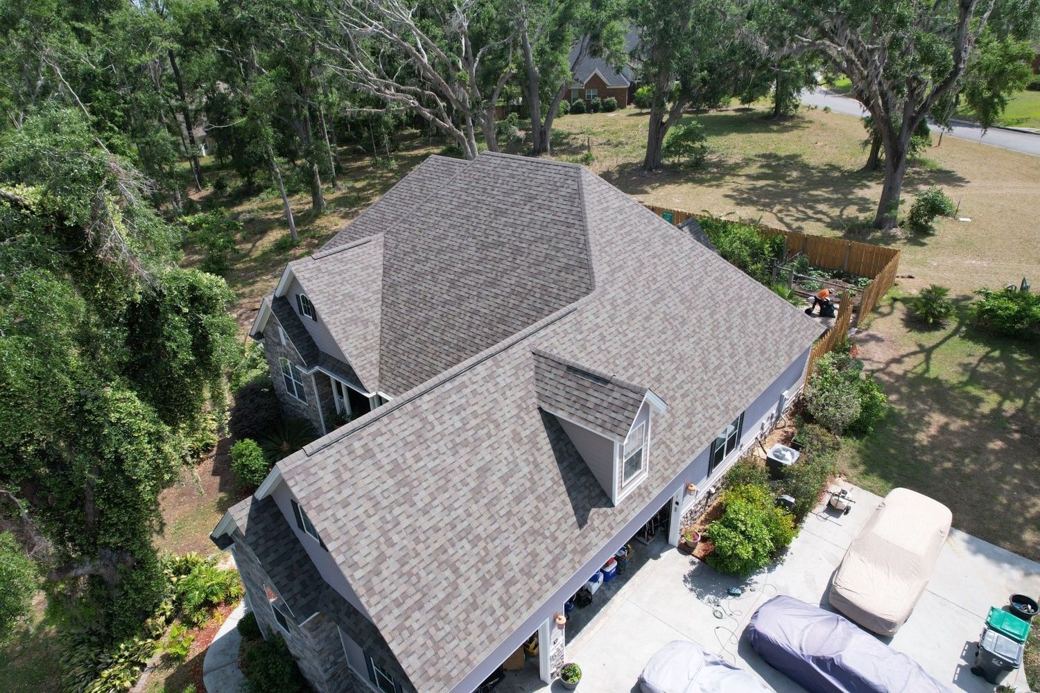 Aerial view of a gray-roofed house with lush green trees surrounding it, driveway with covered cars.
