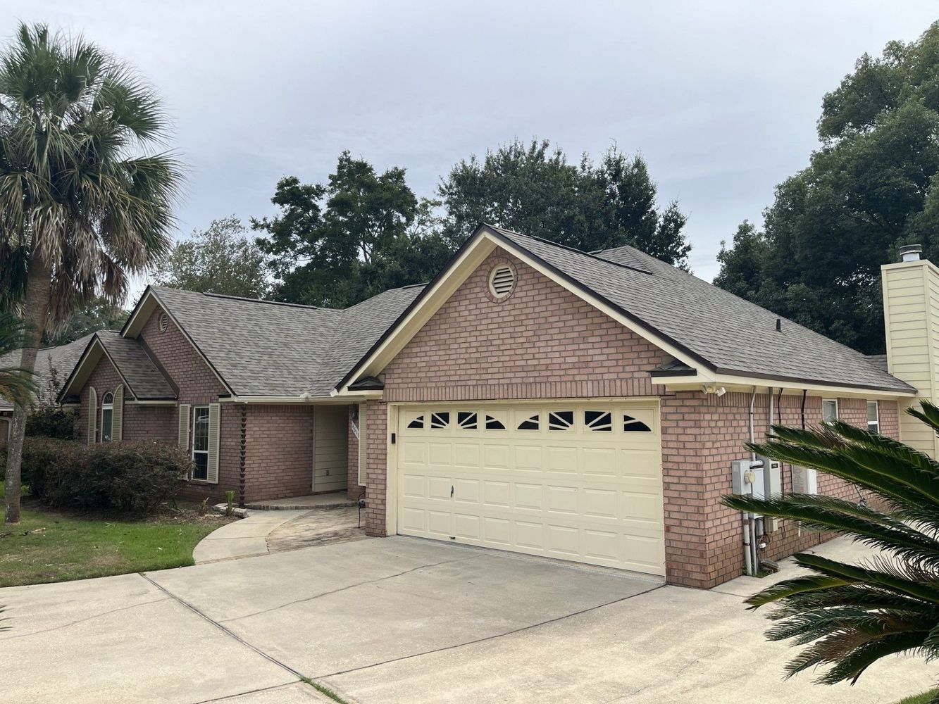 Brick house with gray roof, beige garage door, and a concrete driveway. Trees in background.