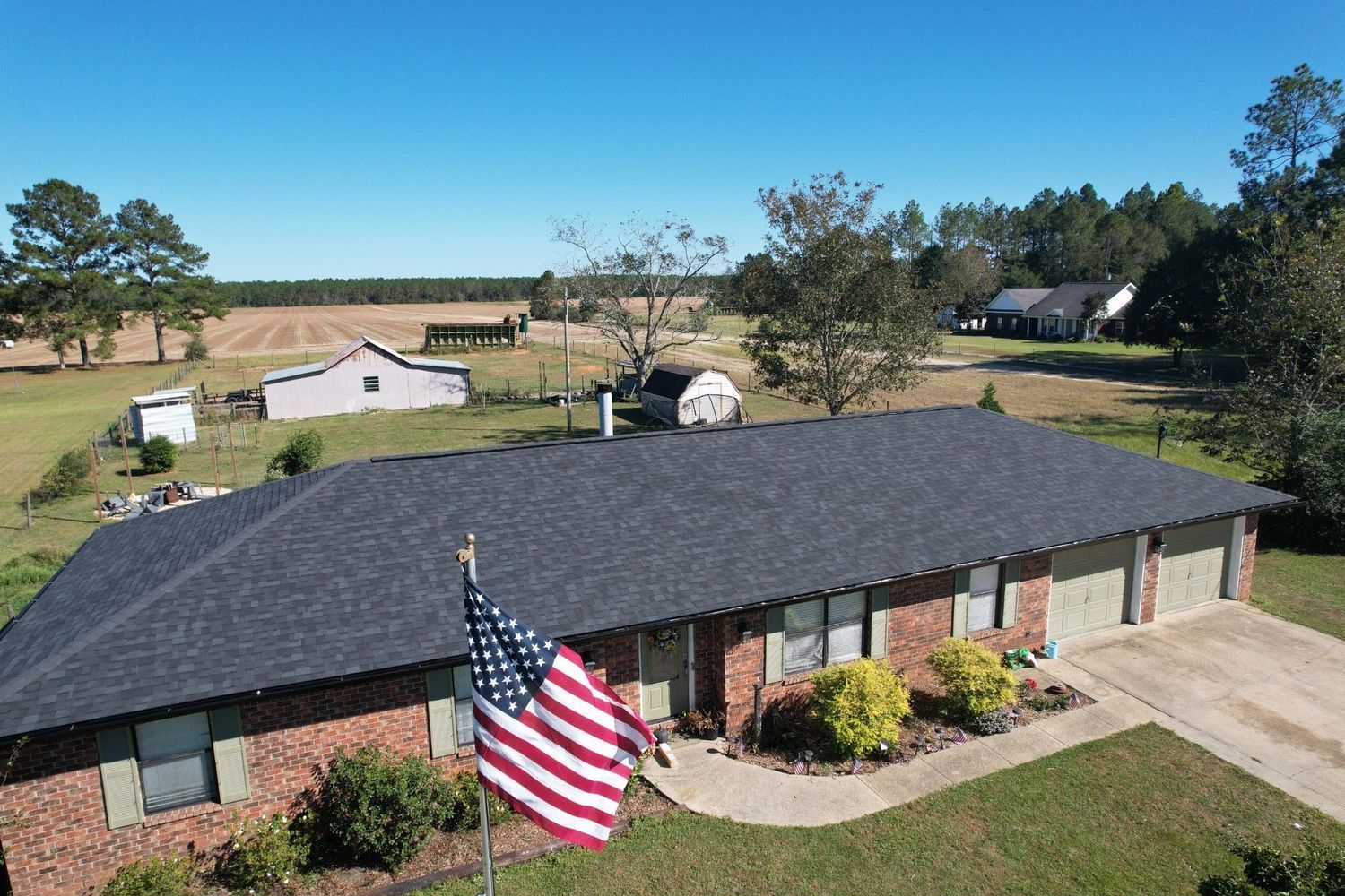 An aerial view of a brick house with a dark roof and an American flag in front, fields and trees in the background.