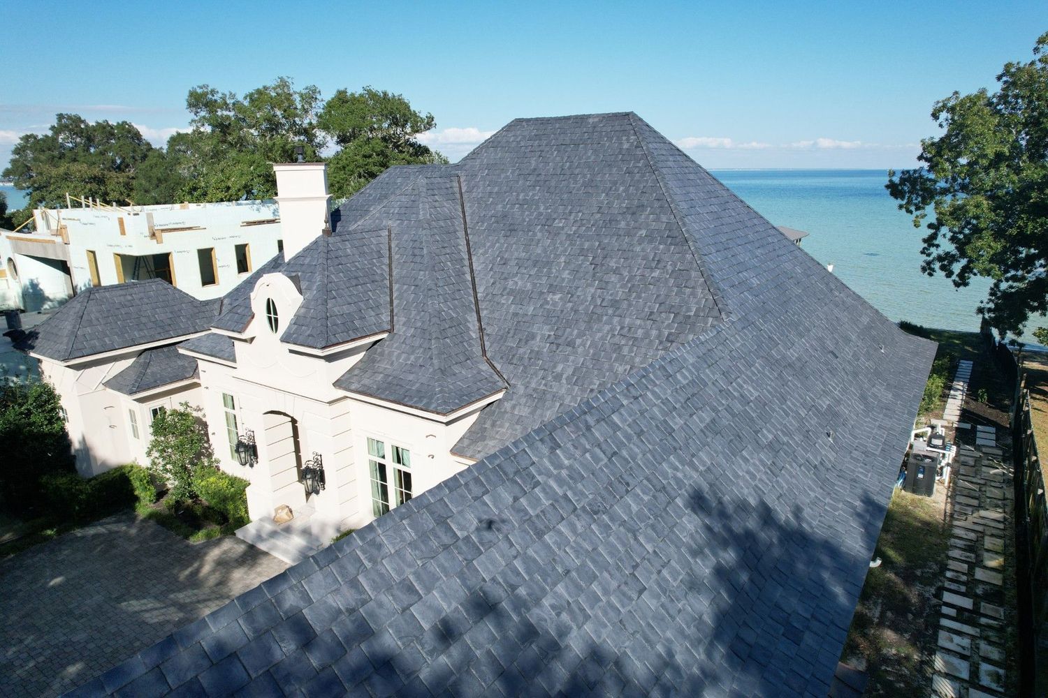 Aerial view of a light-colored brick house with a dark blue shingle roof next to a lake.