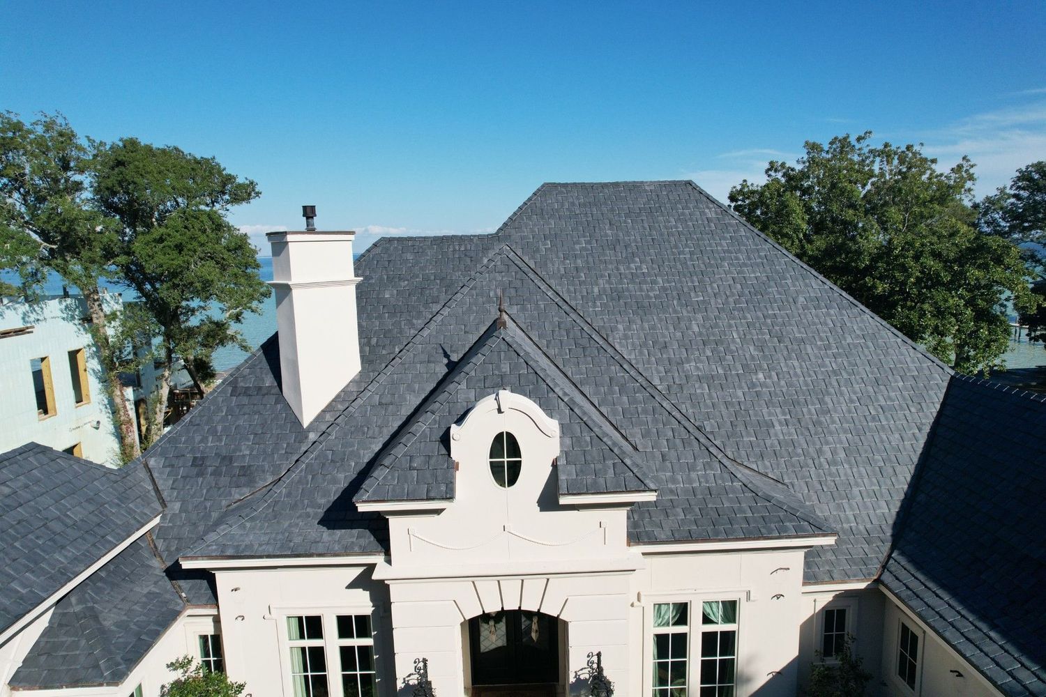 A light-colored stone house with a dark gray roof, chimney, and trees against a blue sky.