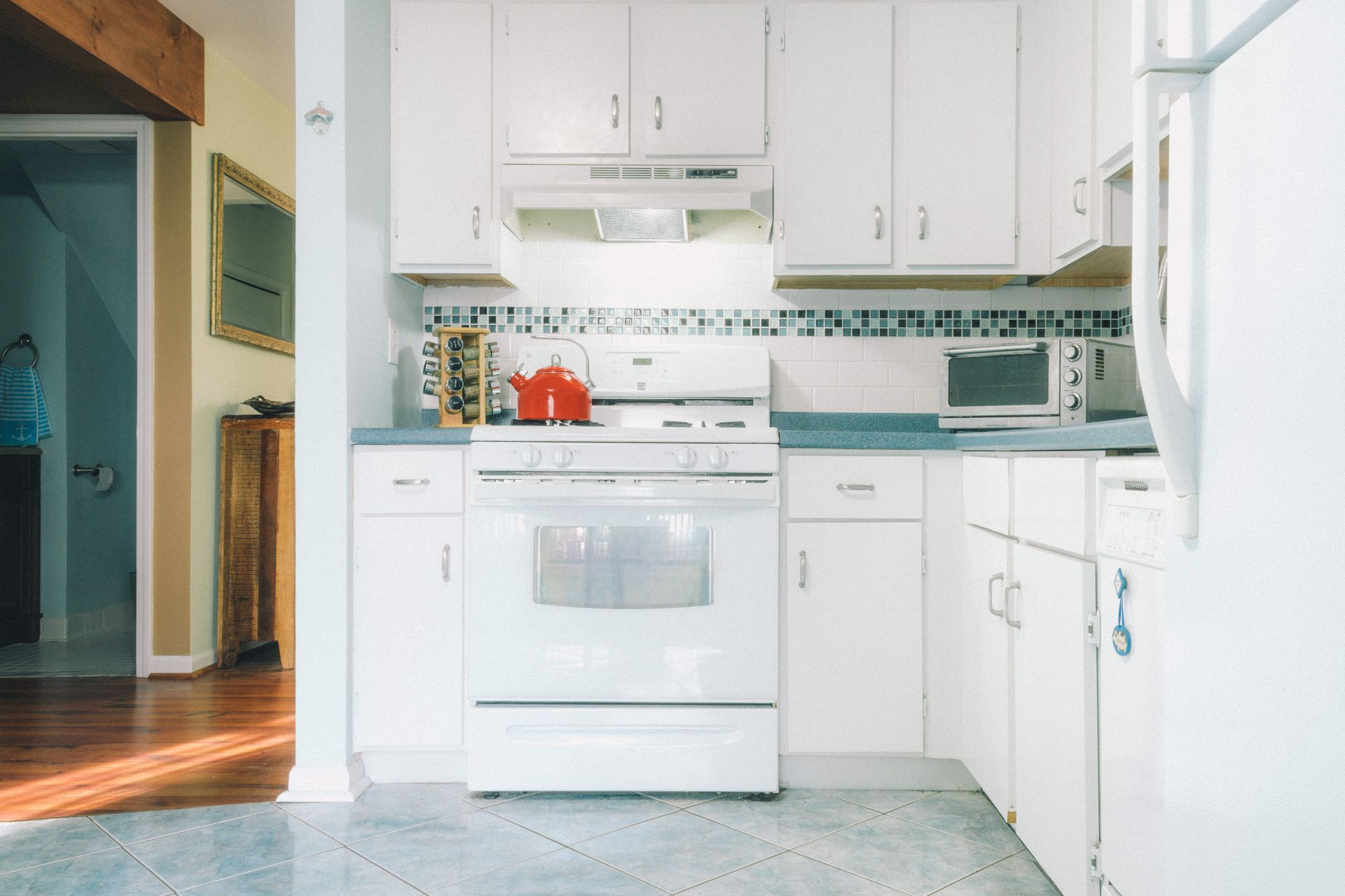 White kitchen with stove, cabinets, and blue countertop. Red kettle on stove.