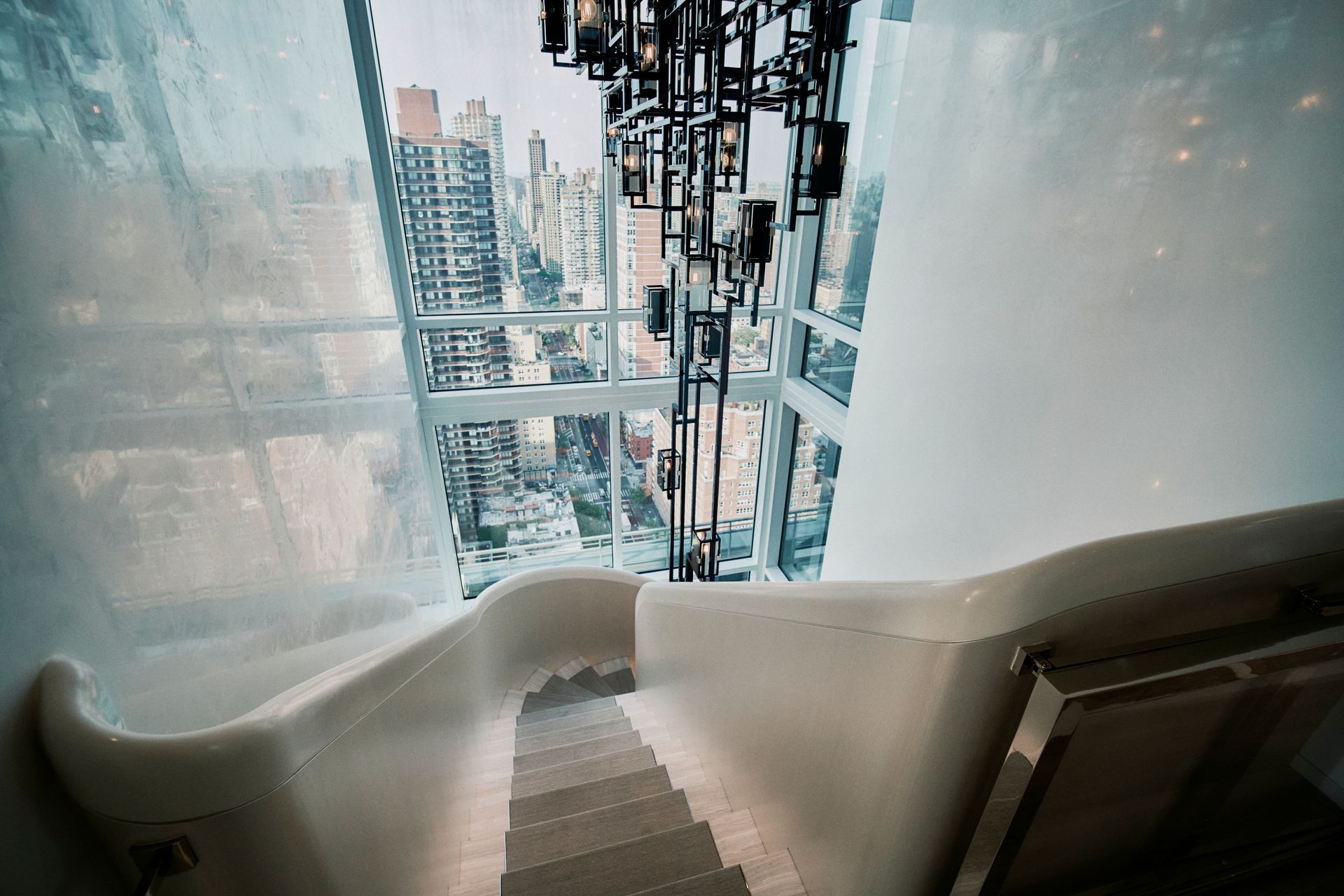 Staircase descending with city view through floor-to-ceiling windows. Modern design with white walls, dark chandelier.