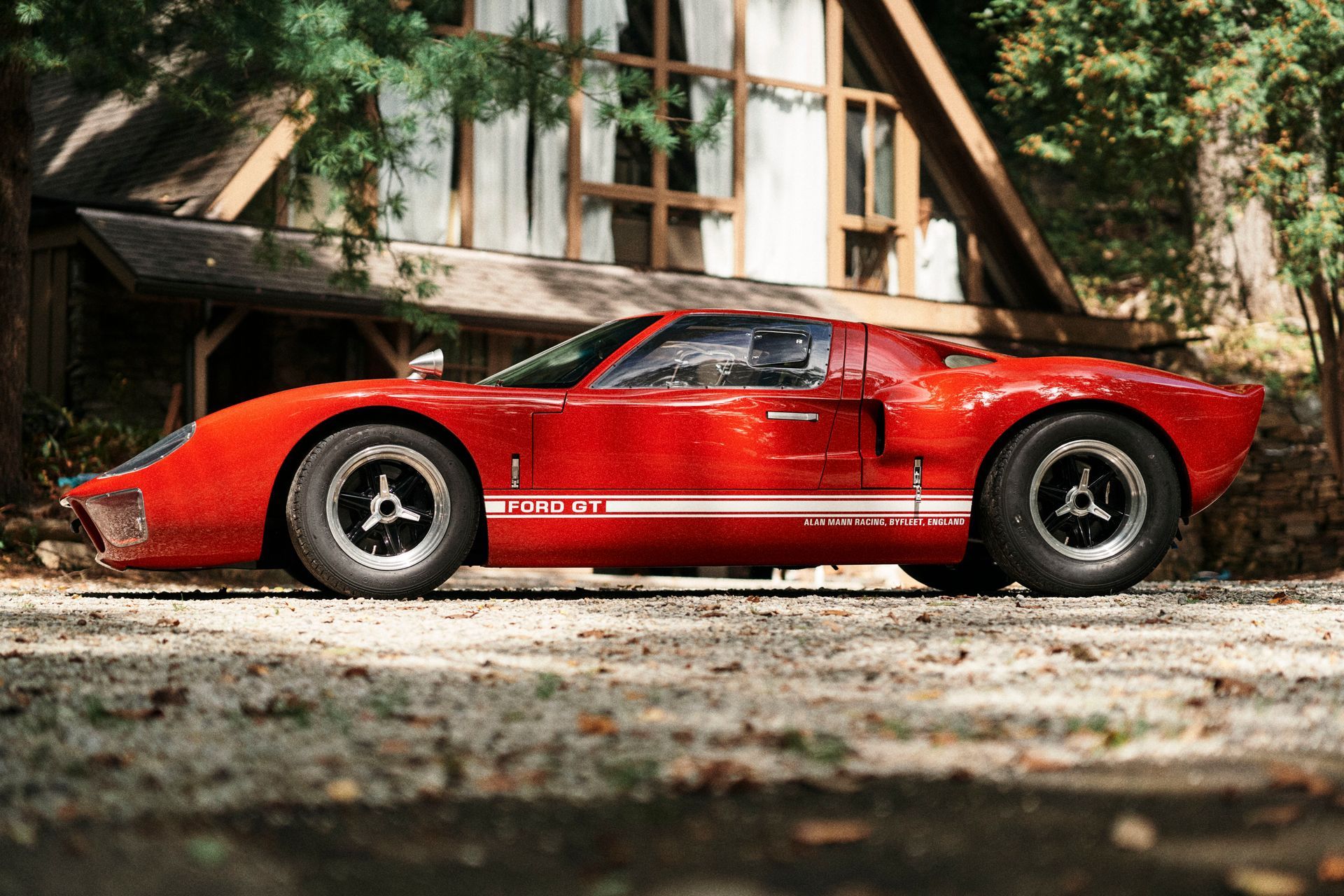 Red Ford GT40 parked in front of a wooden cabin. White racing stripe, black wheels, and a sunny day.