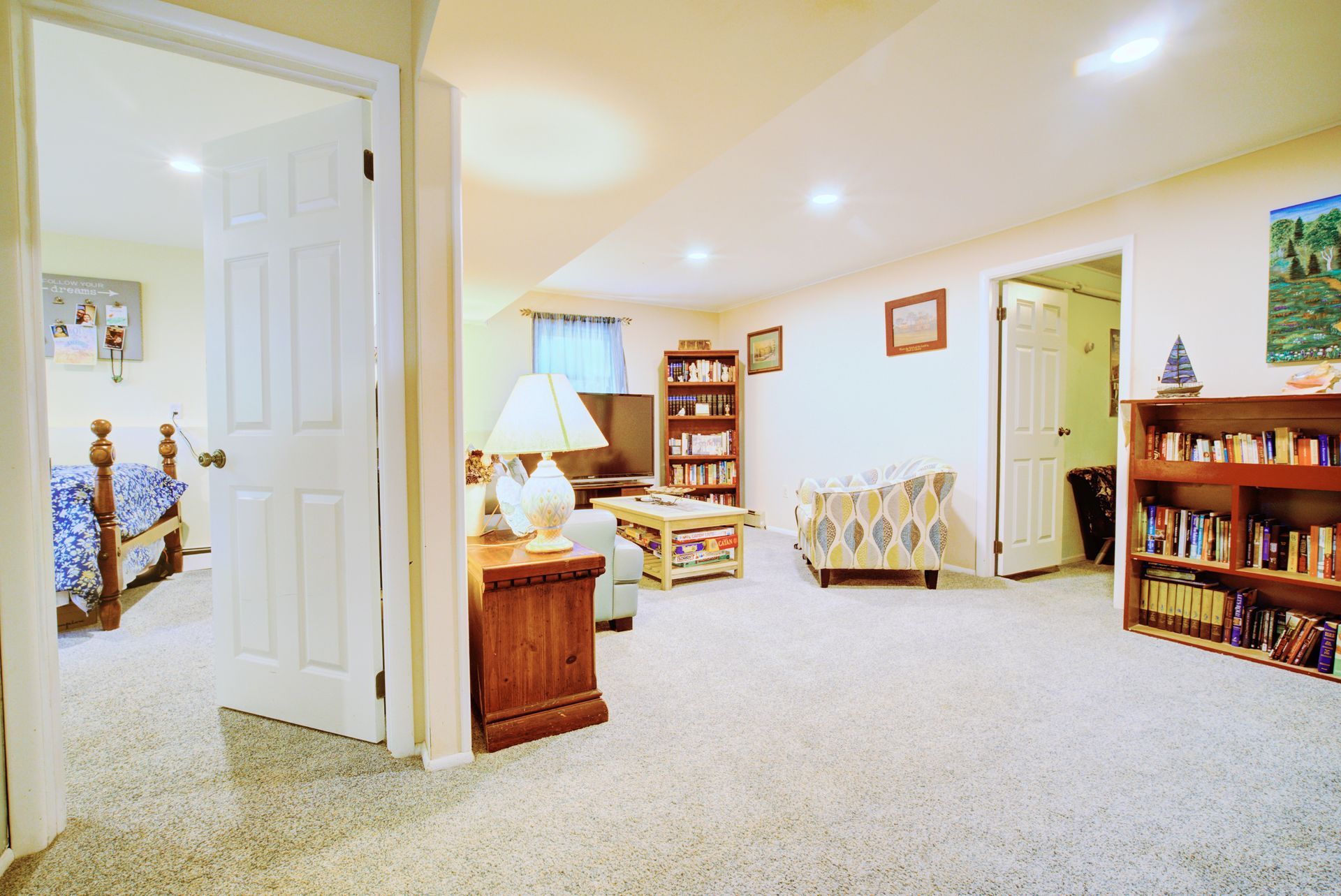 Cozy basement living room with beige carpet, bookshelves, TV, and open doorways to bedrooms.