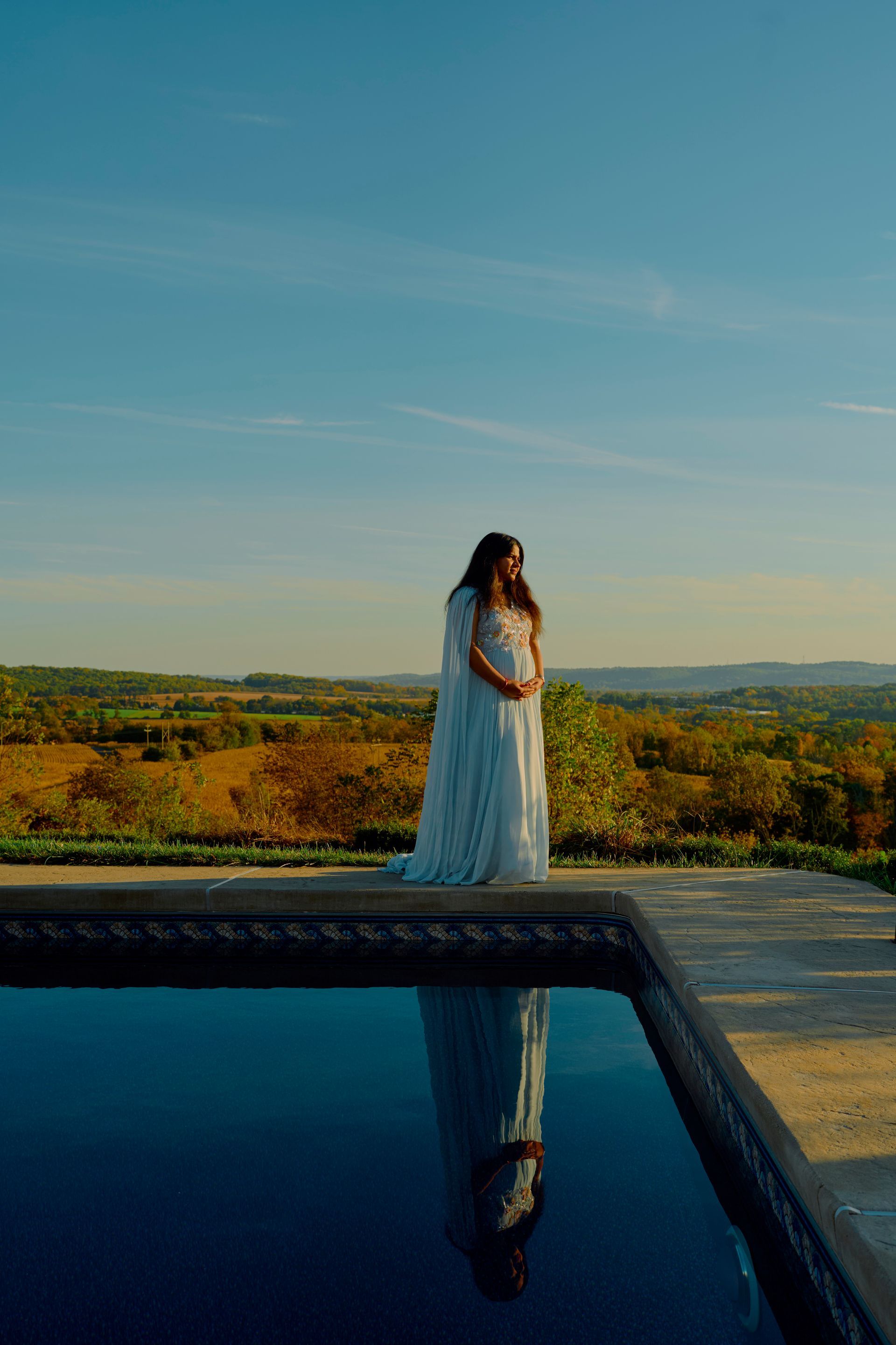 Pregnant woman in a white dress stands by a pool, overlooking a landscape under a blue sky.