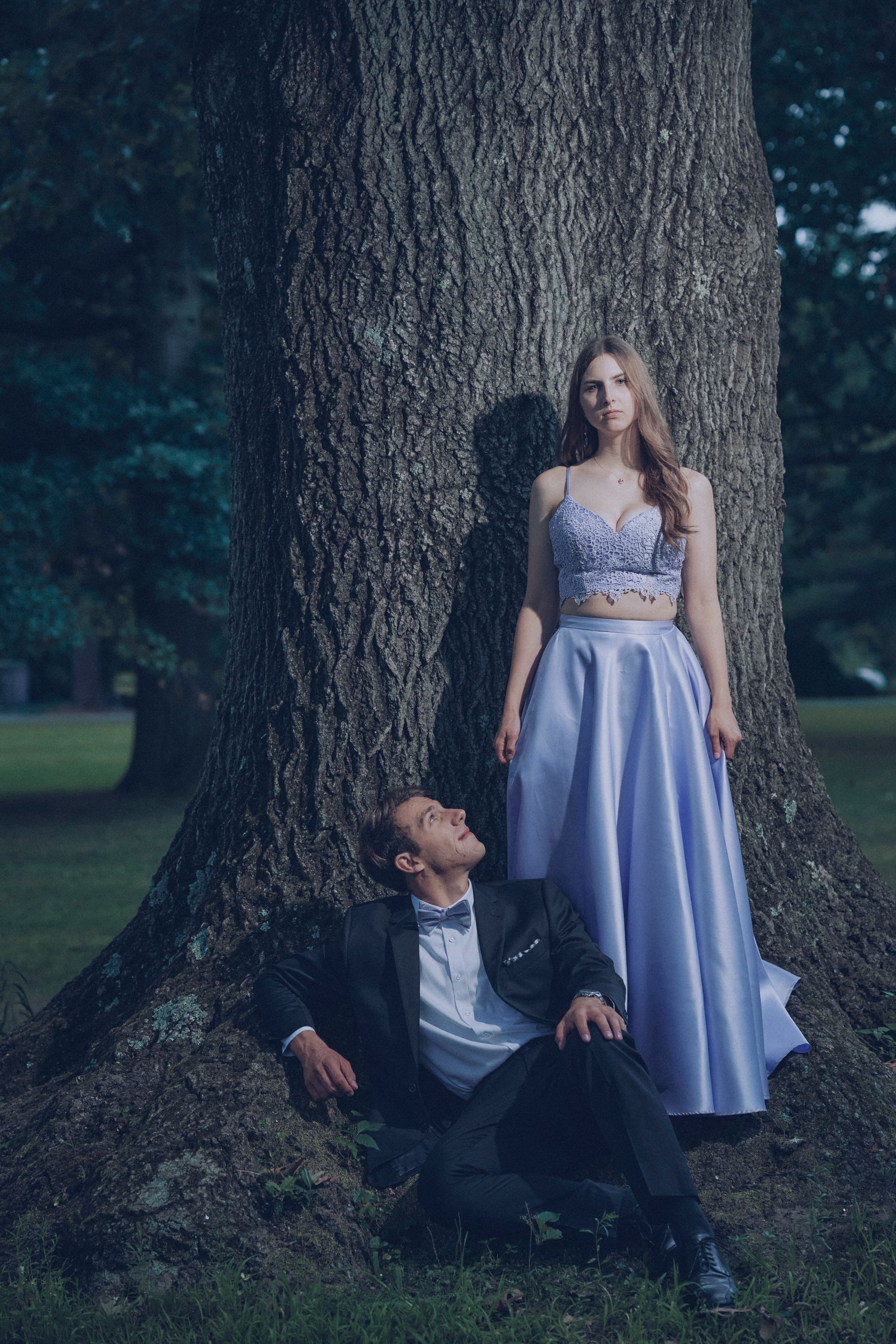 Couple in formal attire, she in lilac gown, he in tuxedo, near a tree.