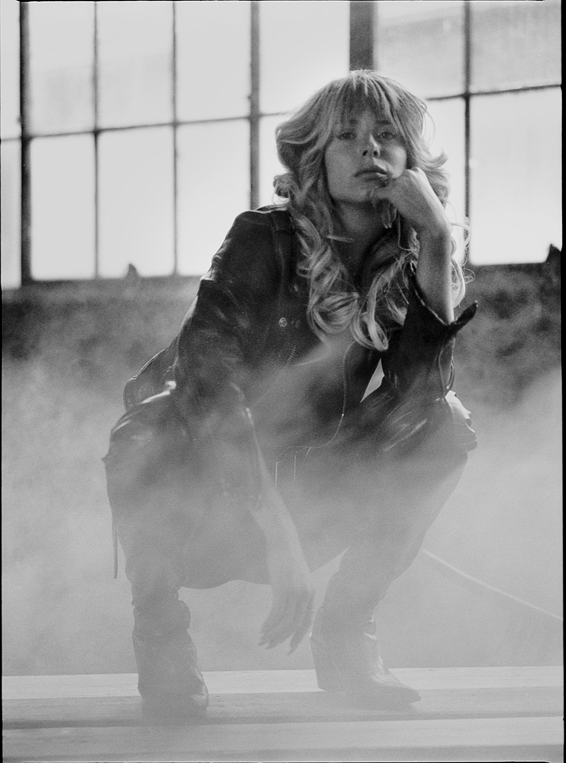 Blond woman, leather jacket, crouches, hand on chin, amid dust in industrial setting, gazing thoughtfully.