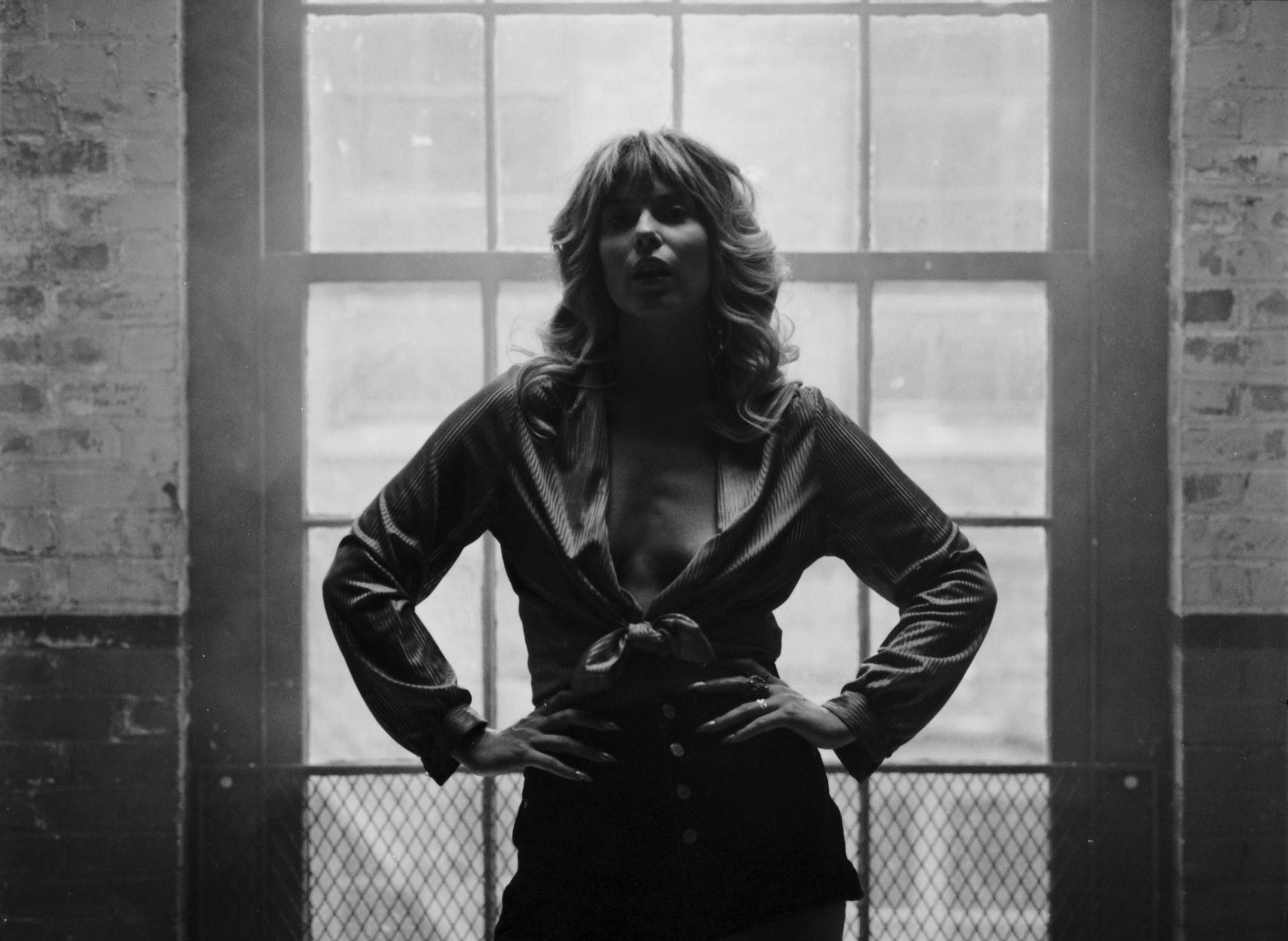 Woman in low-cut shirt, hands on hips, stands in front of a window, in black and white.