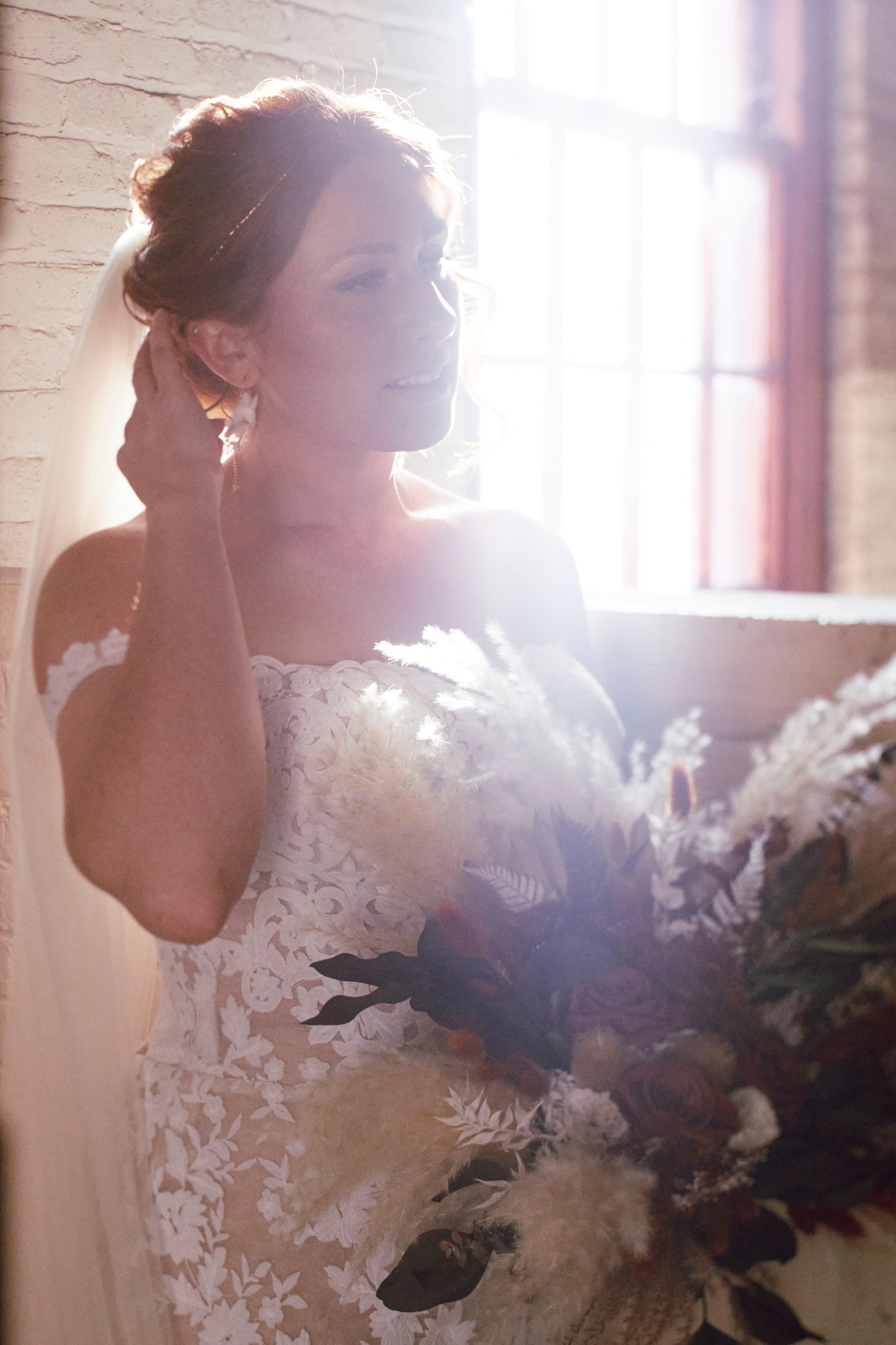 Bride in lace dress, holding bouquet, adjusts her earring in sunlight.