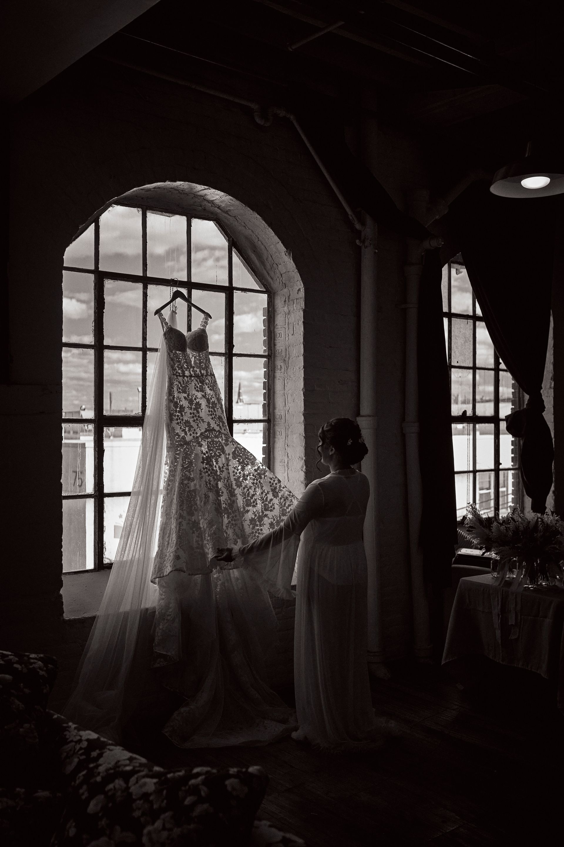 Wedding dress hanging in a window, woman touching the gown. Black and white interior setting.